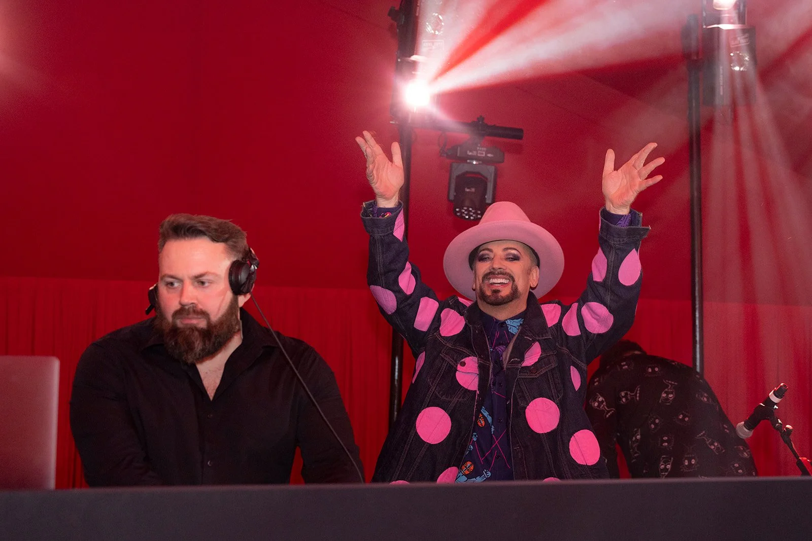 Two men are on a red stage; one man wearing a black shirt with headphones appears serious, while the other man in a pink polka-dot jacket, pink hat, and vibrant makeup is smiling and raising his arms with hands open.