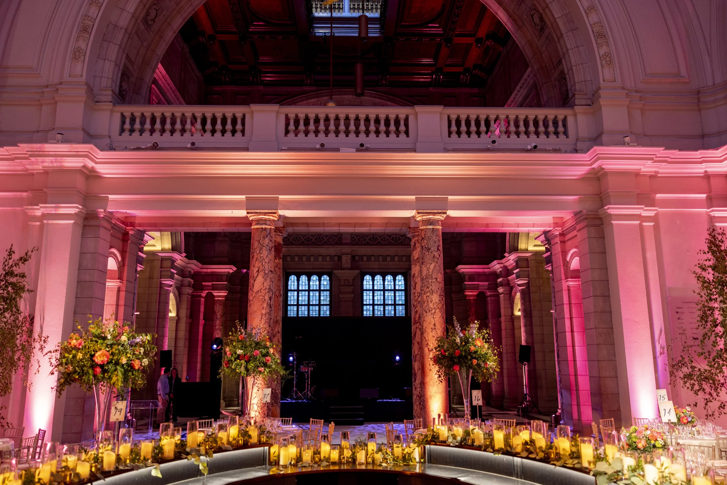 Interior of a grand hall decorated with pink lighting, tall marble columns, floral arrangements, and tables set for a formal event.