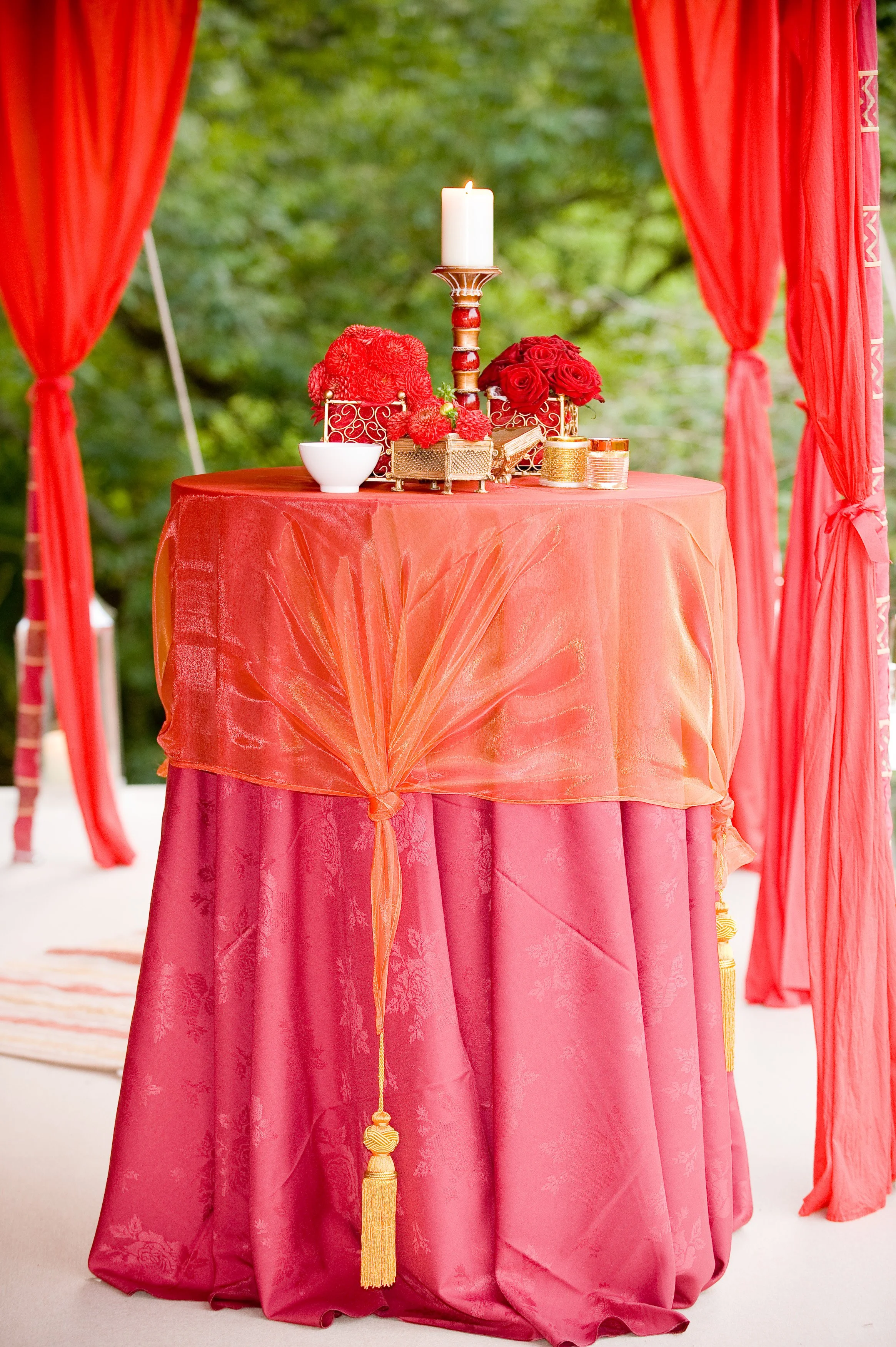 A round table draped with a pink satin tablecloth featuring floral patterns, set up outdoors with red curtains on either side. The table has a gold finish candlestick with a white candle, arranged red roses, a small white bowl, gold decorative contai