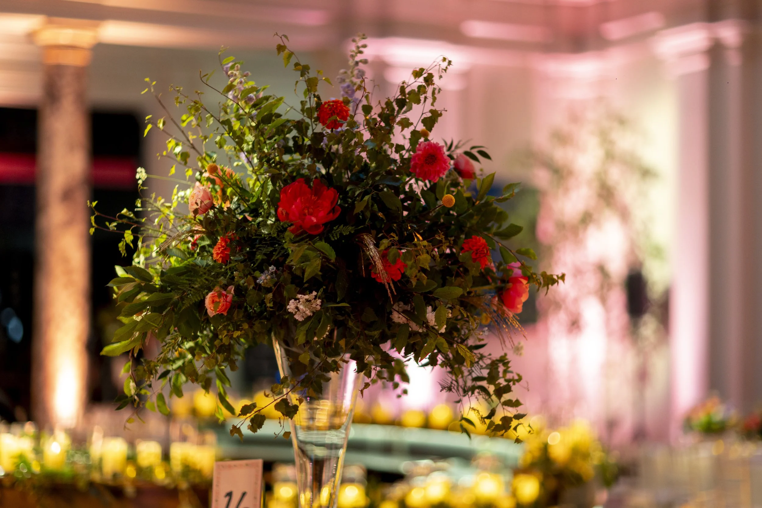 A bouquet of various colorful flowers arranged in a tall glass vase on display at a flower market with warm lighting in the background.