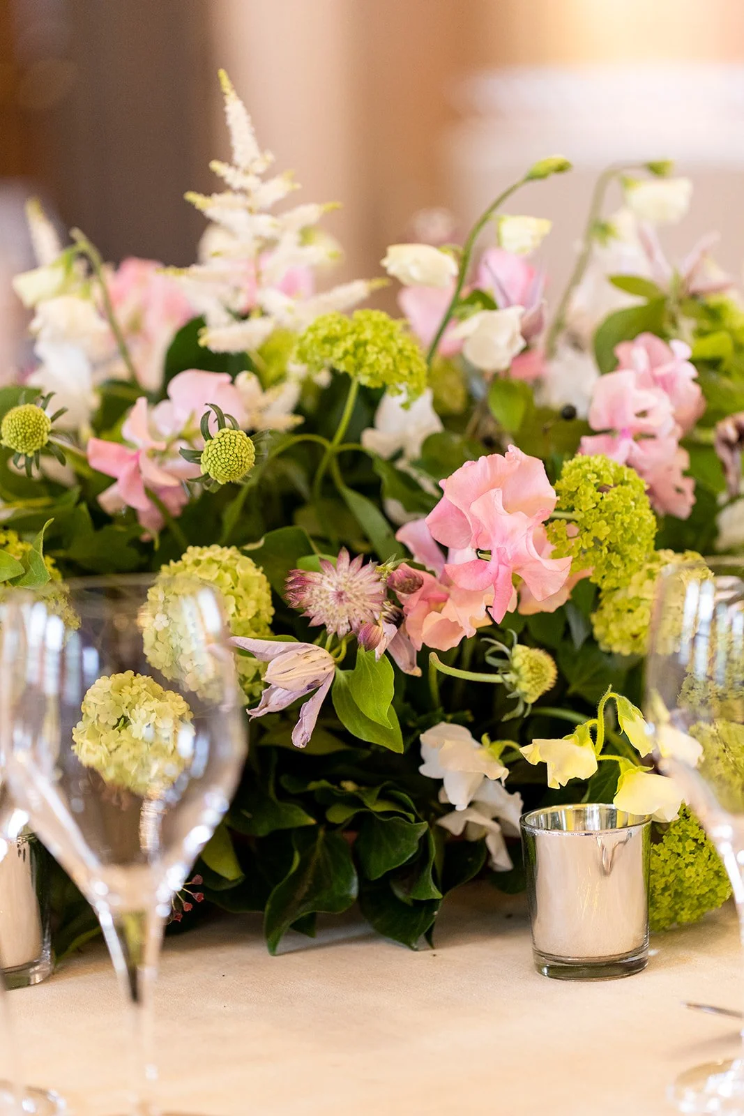 Close-up of a floral arrangement with pink, white, and green flowers on a table, surrounded by wine glasses and a candle in a glass holder.