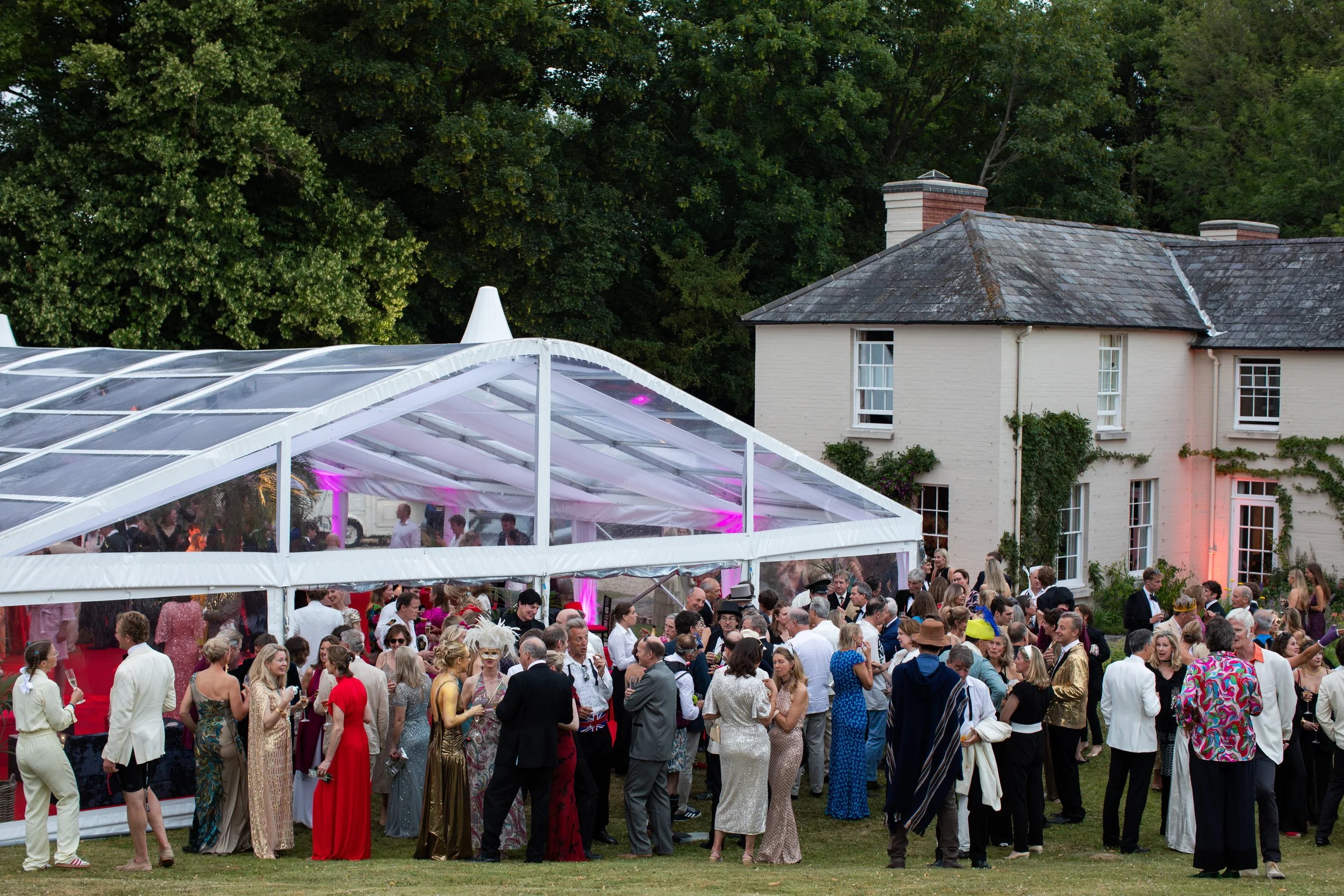 A large outdoor party with many people dressed in elegant and colorful clothing, gathered under a clear tent on a grassy area, next to a white brick house with green foliage and tall trees in the background.