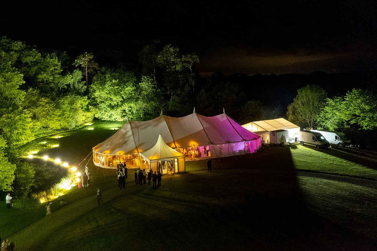 Nighttime outdoor event with illuminated large white tent and smaller attached tents, surrounded by trees and grass, with a few people gathered near the entrance and parked vans nearby.