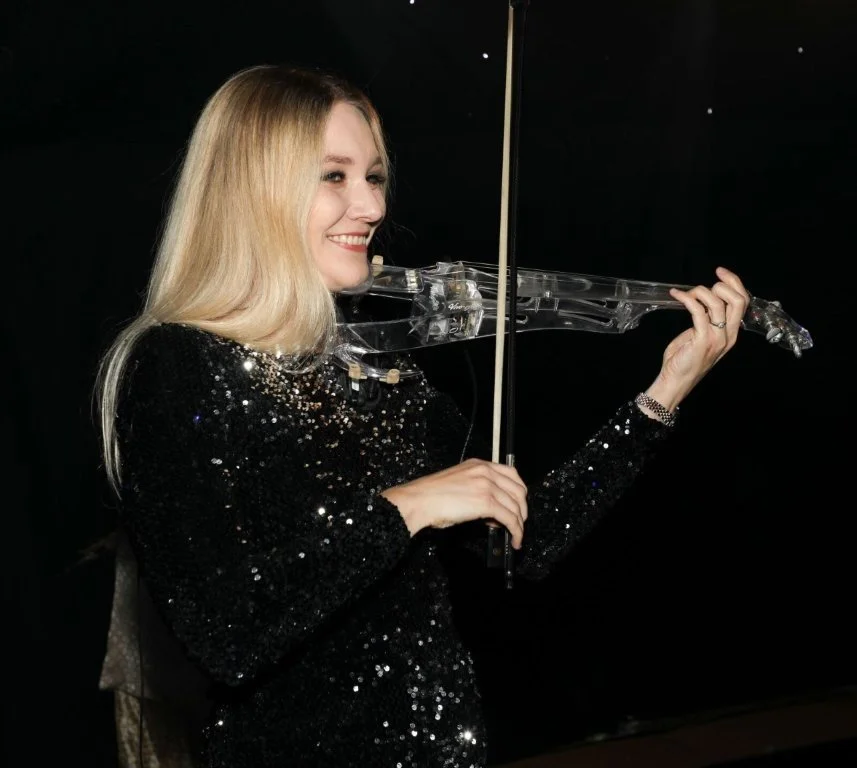 A woman with blonde hair playing a transparent electric violin, smiling, wearing a black sequined dress, against a black background.