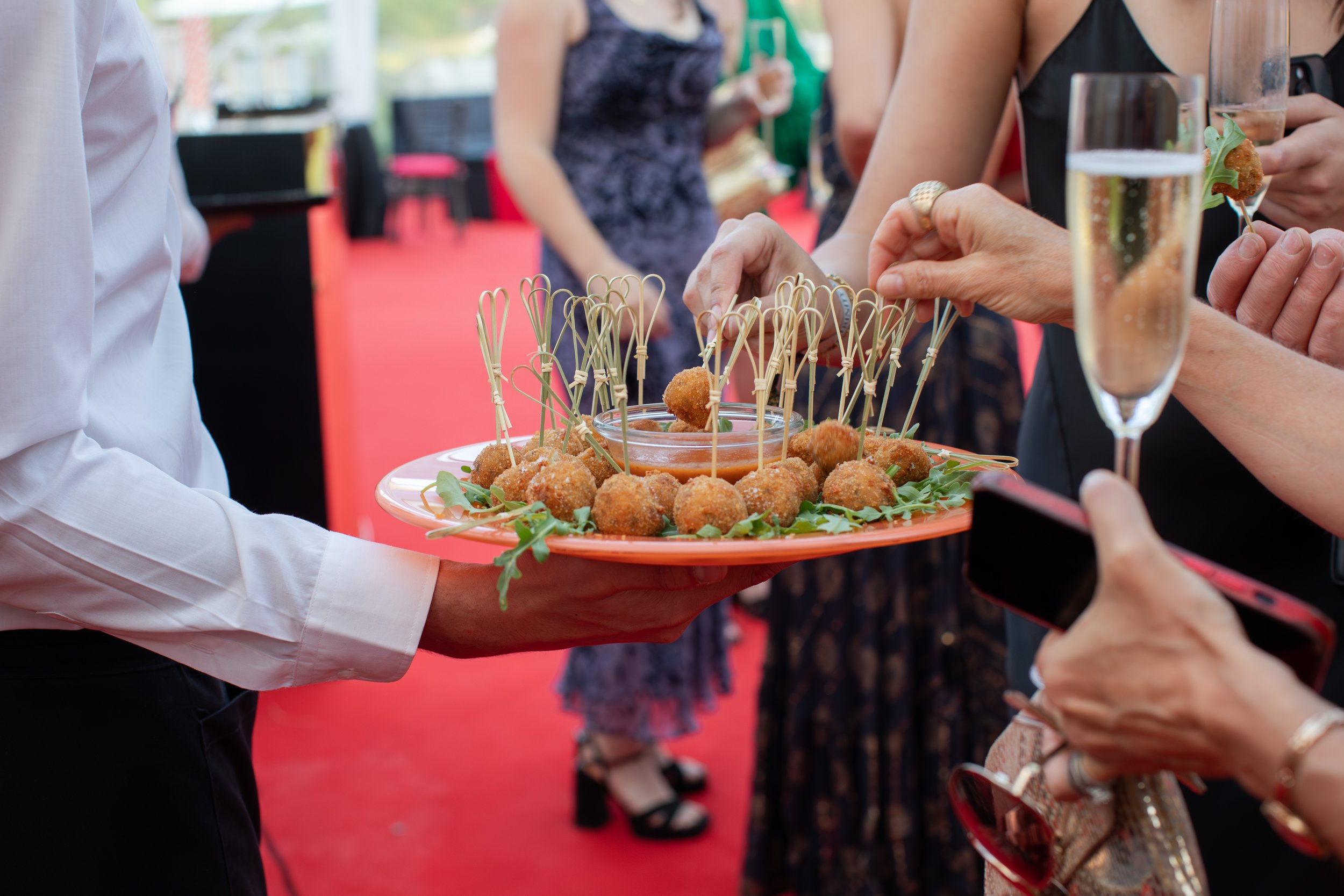 Person serving a platter of breaded appetizers with skewers to guests at an indoor event.