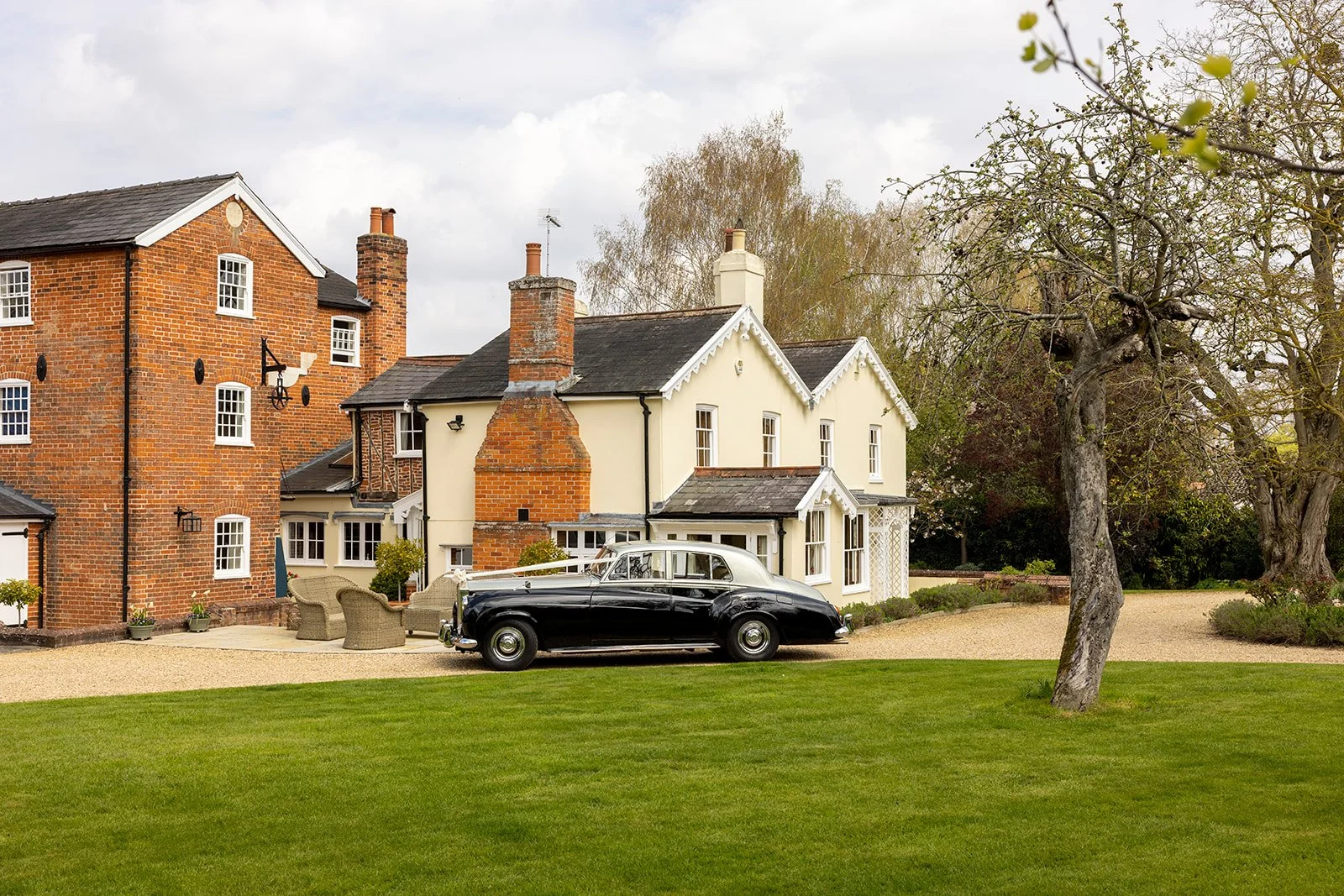 A vintage black and white car parked on a gravel driveway in front of a large traditional house with a white facade and red brick chimneys, surrounded by green lawn and trees.