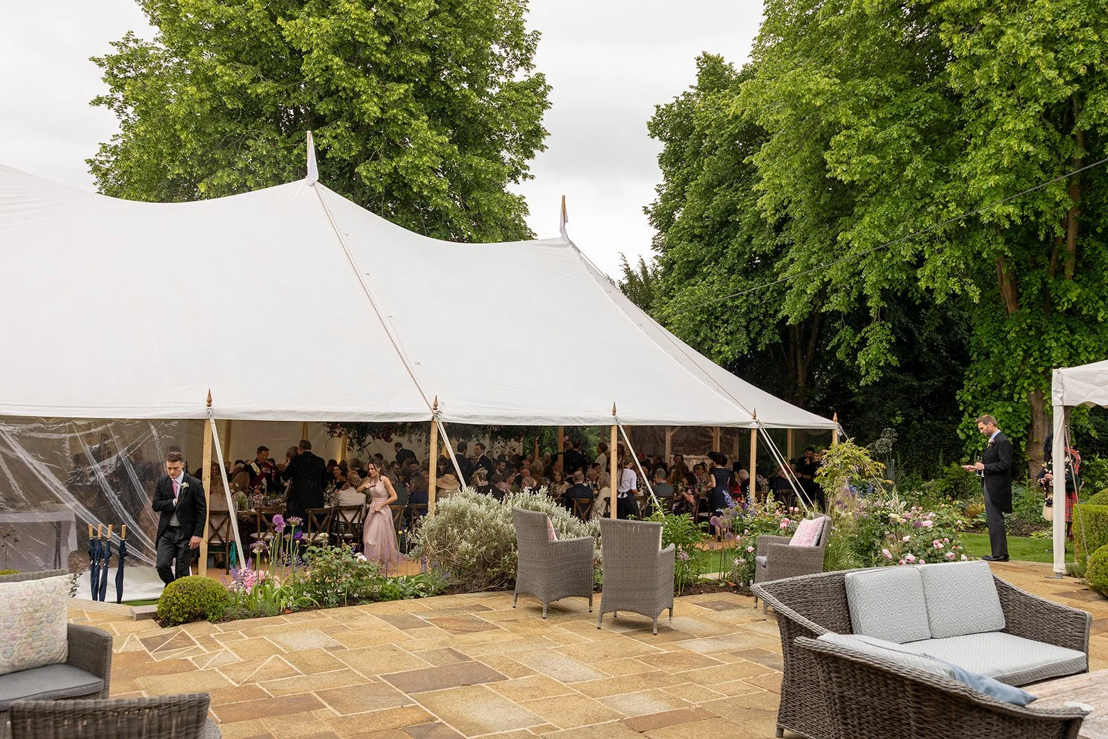 A wedding reception under a large white tent in a garden with trees. Guests are seated at tables inside the tent, while some are standing outside. There are chairs, sofas, and potted plants arranged on the brick patio.