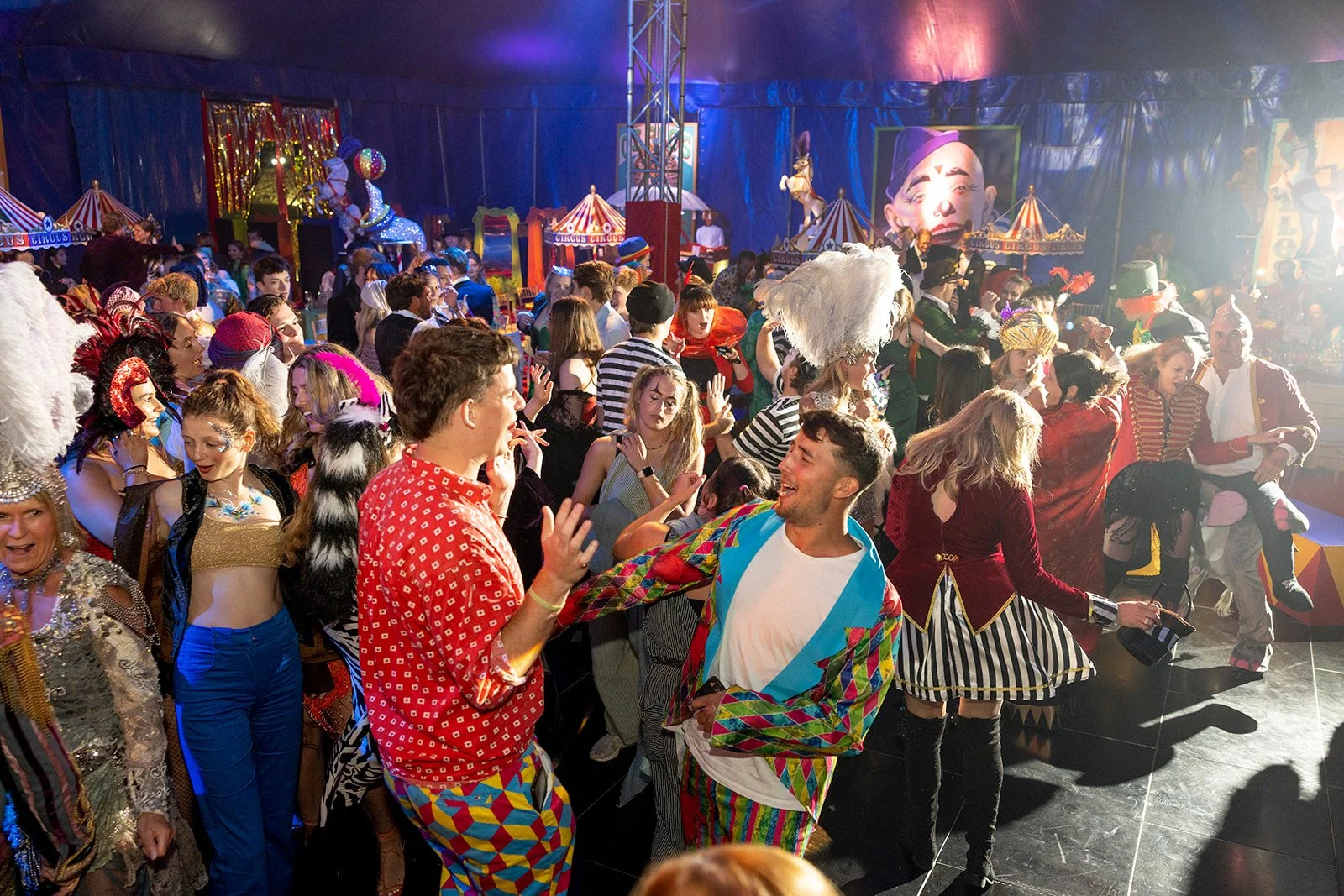 People dressed in colorful costumes and vintage circus outfits dancing and enjoying a carnival or circus-themed party in a decorated tent with carnival rides and large clown face poster in the background.