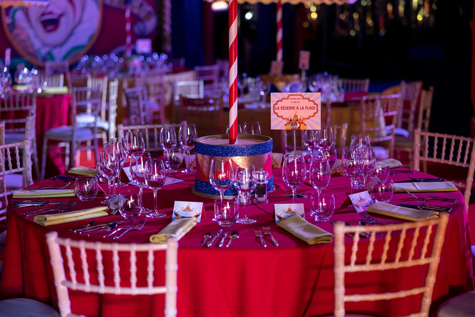 A round banquet table decorated with a red tablecloth, set with multiple wine glasses, champagne flutes, and silverware, with yellow napkins folded on the plates. The table has a centerpiece designed like a carousel with a red and white striped straw