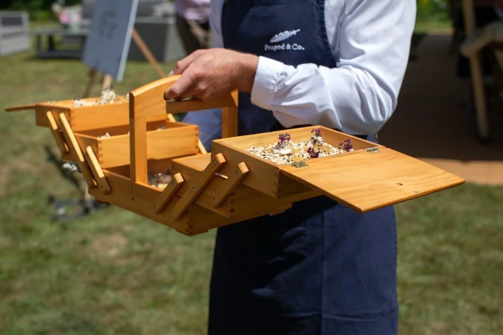Person holding a wooden tray with compartments containing small rocks and flowers.
