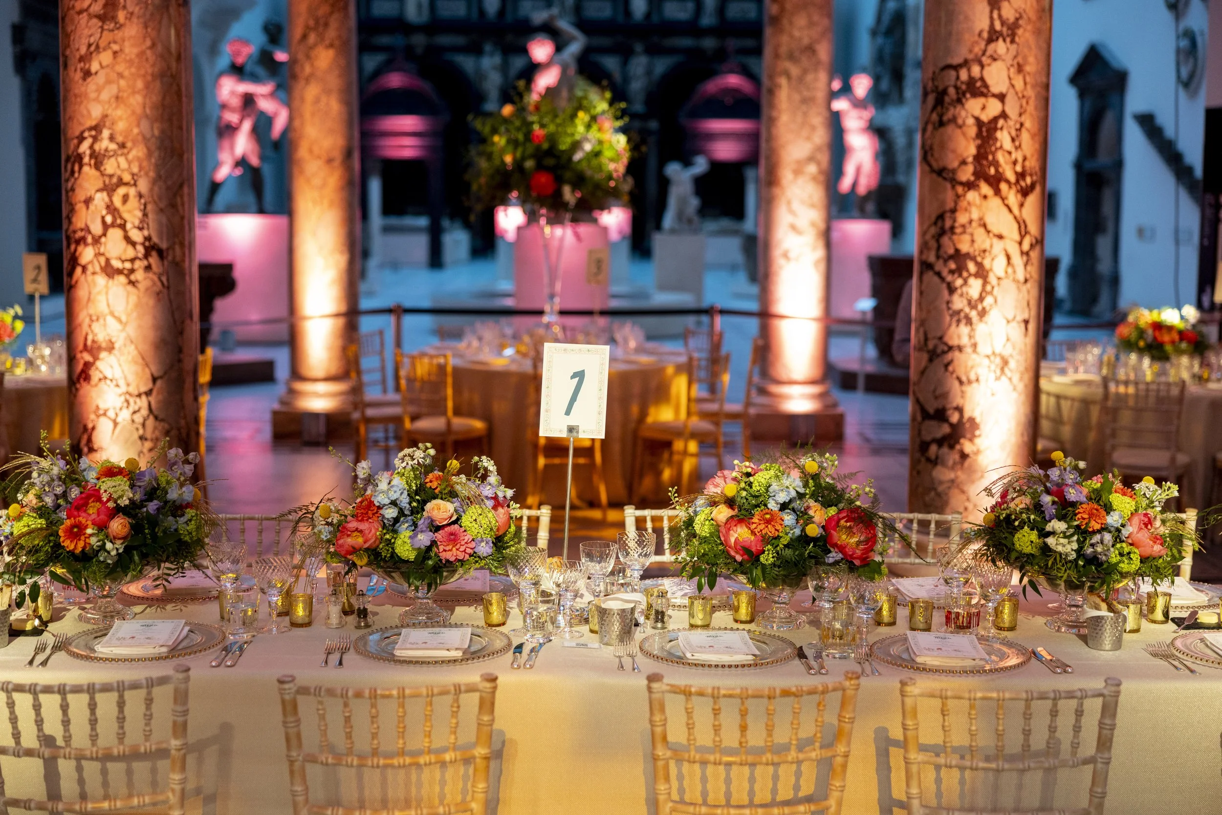 Elegant banquet table set for a formal event with floral centerpieces, candles, and place settings, in a decorated venue with large columns and artwork.