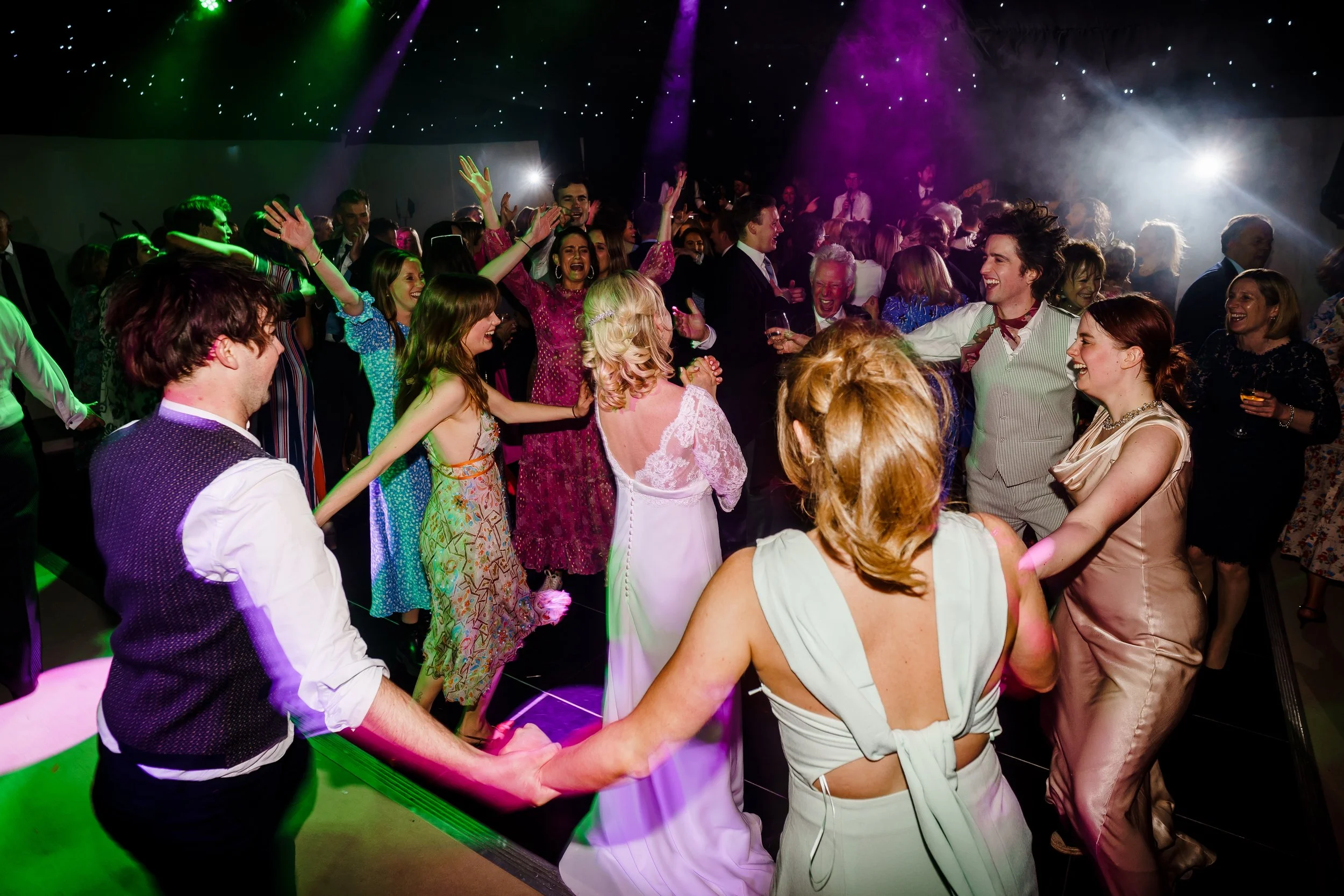 People dancing and celebrating at a lively party with colorful lighting, some holding hands and smiling.