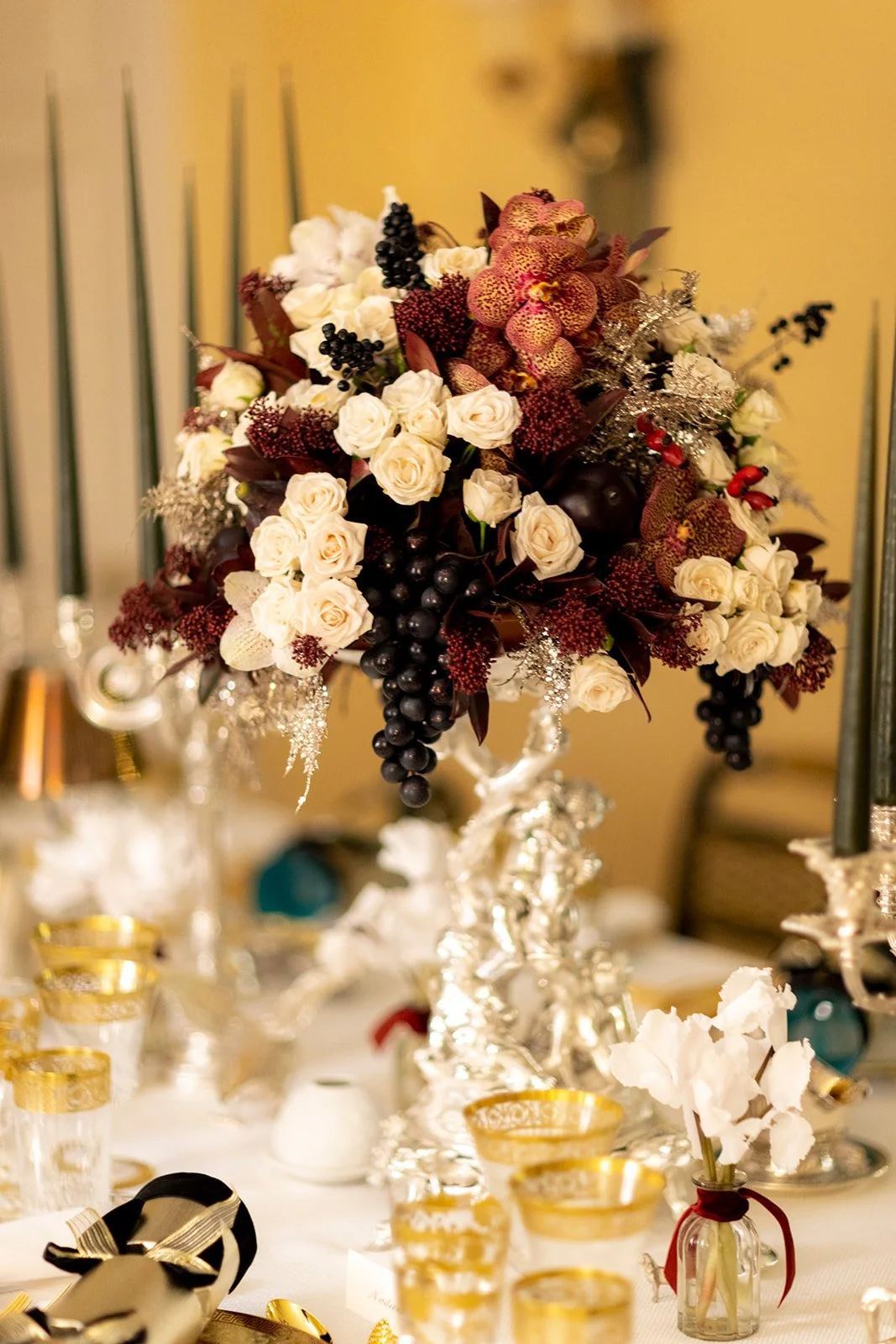 A floral centerpiece with white roses, purple berries, orchids, and dark leaves on a silver ornate stand, arranged on a decorated table with gold-rimmed glasses and small white flowers.