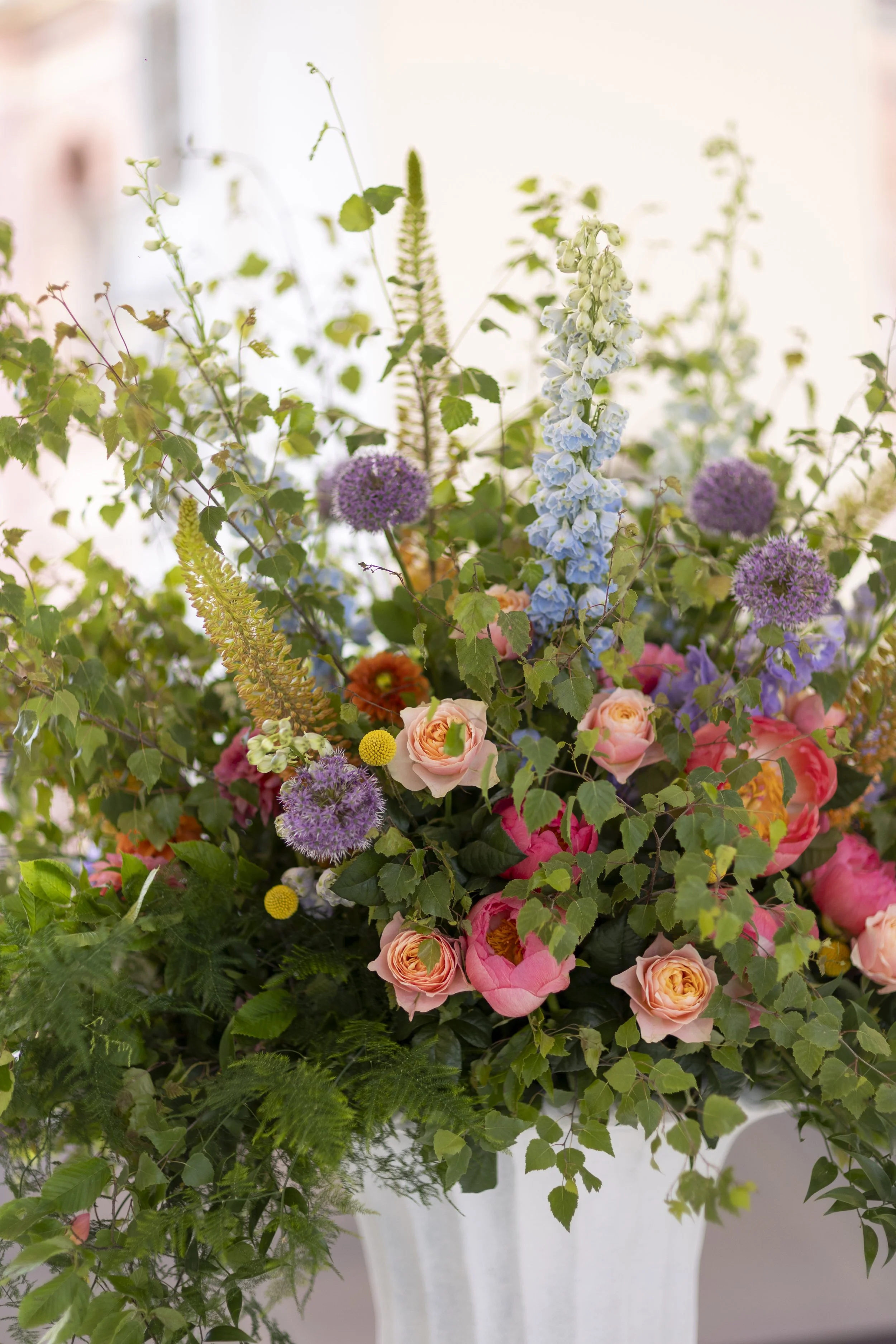 A bouquet of mixed flowers including pink roses, purple alliums, blue delphiniums, yellow billy balls, and various green foliage in a white vase.
