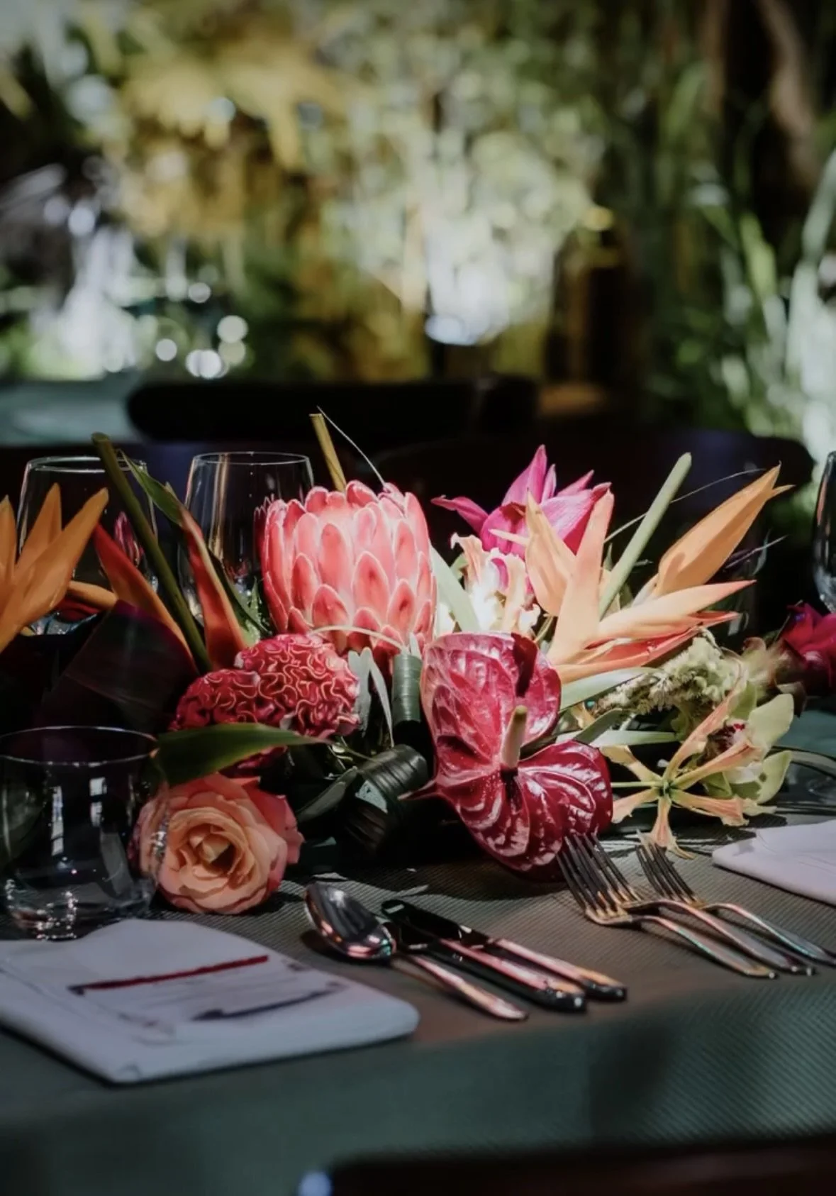 Table setting with a large colorful floral centerpiece featuring pink, red, and orange flowers in a dark green setting.