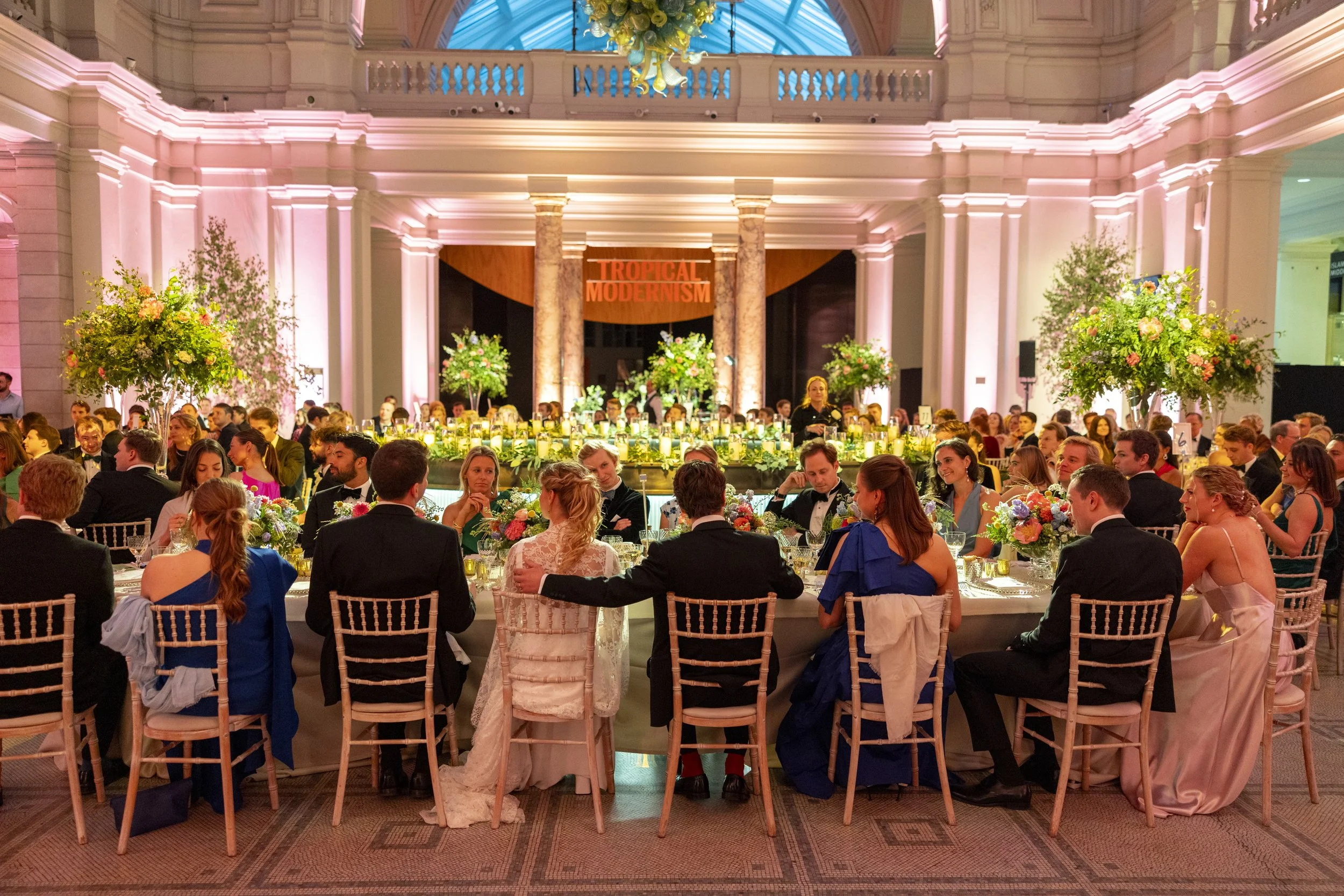 Elegant banquet hall with high ceilings, illuminated pink lighting, and large floral arrangements on tables. A sign reads 'Tropical Modernism' in the background, with guests dressed in formal attire seated around the tables.