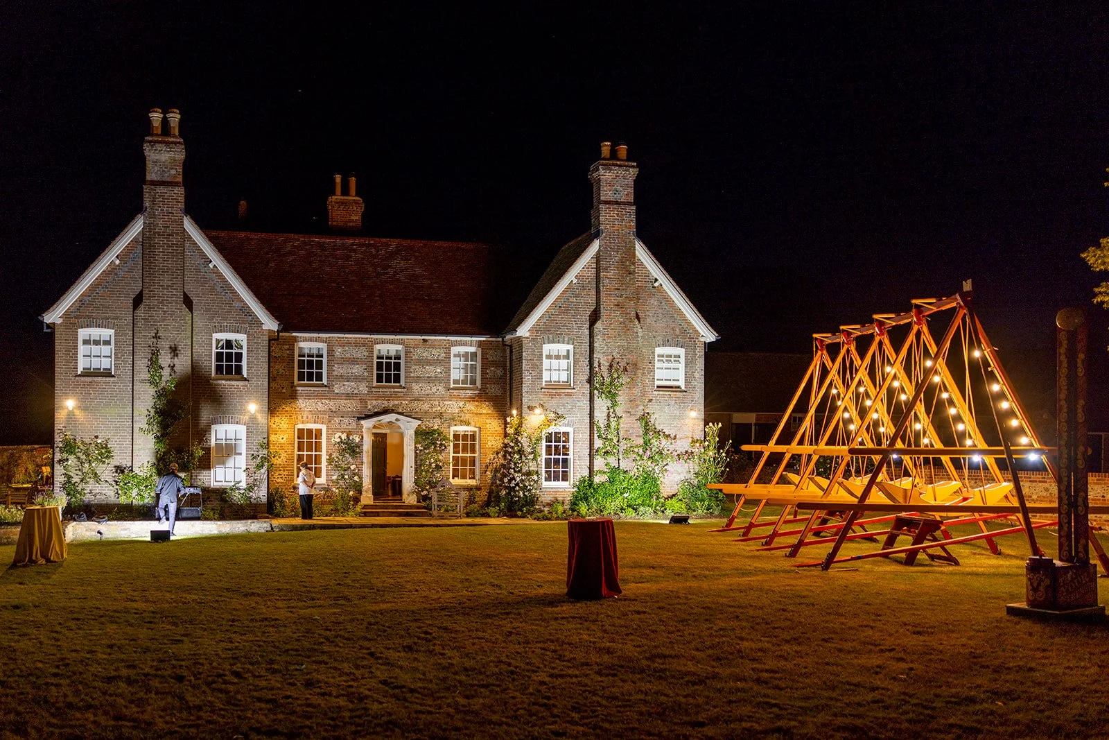 Night view of a brick house with lit lanterns and a garden, with a playground set on the right side decorated with lights.