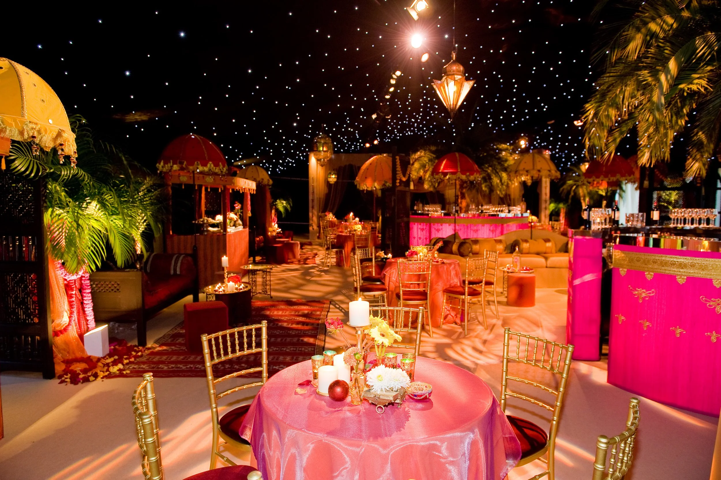An ornate indoor celebration setup with pink and gold decorations, tables with candles, and umbrellas, under a ceiling decorated with hanging lanterns and string lights, creating a festive atmosphere.