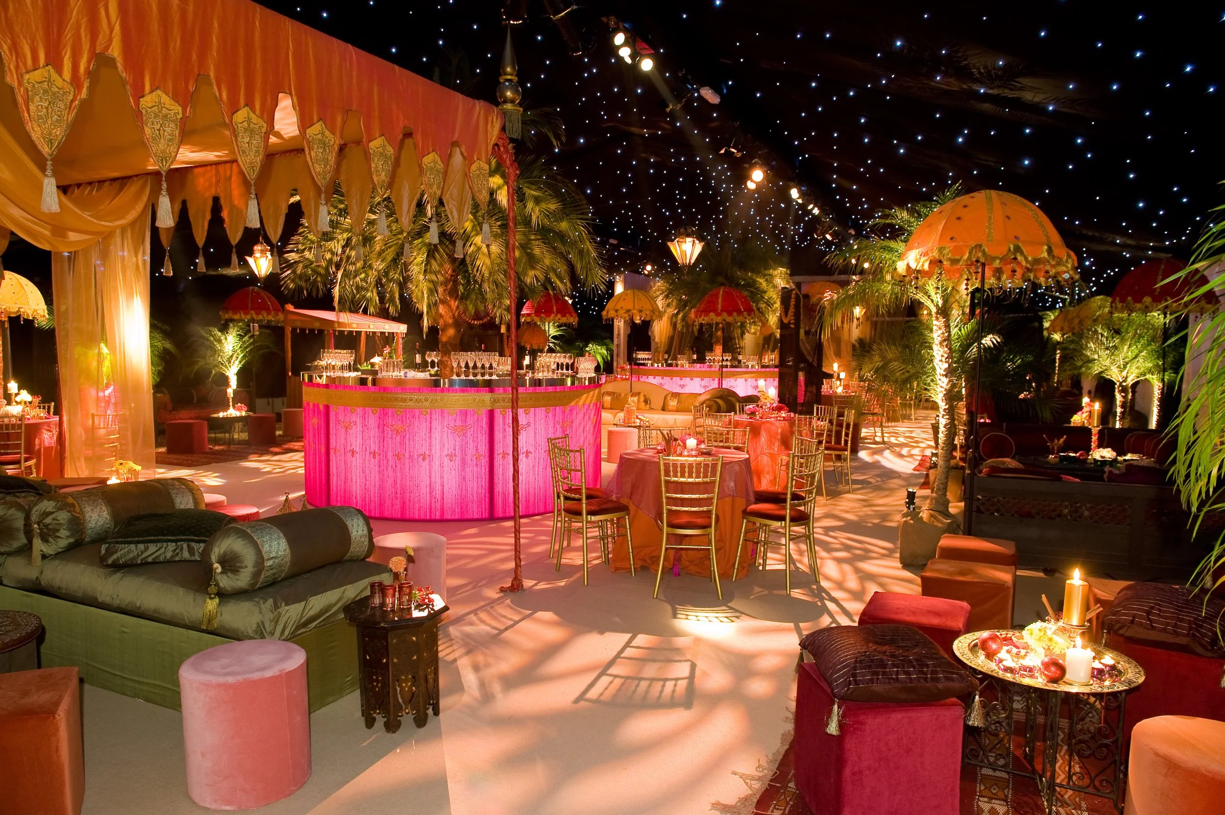 Decorated event space with pink and gold accents, round tables with chairs, velvet sofas, palm trees, and hanging umbrellas under a starry ceiling.