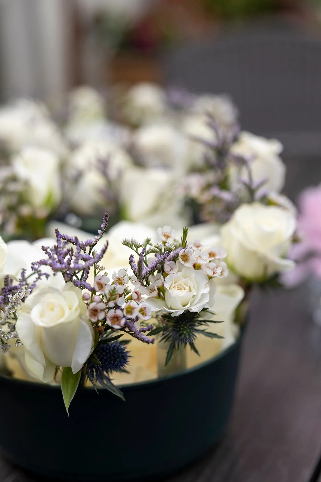A floral arrangement in a black bowl with white roses, small white and pink flowers, purple sprigs, and blue thistle flowers on a dark surface.