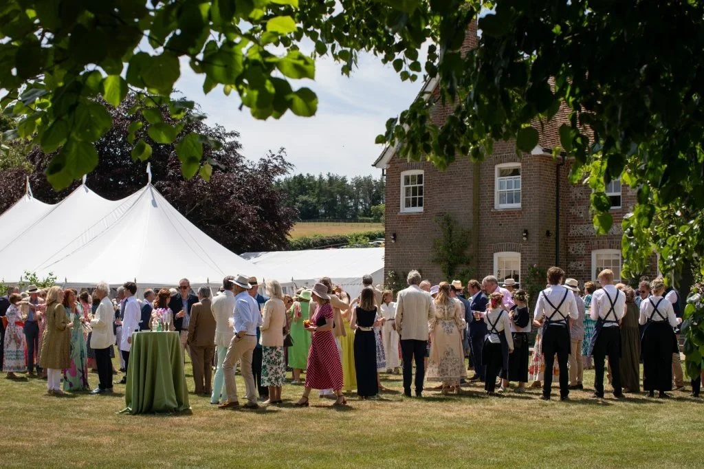 Guests at an outdoor event in vintage attire gather on a grass lawn near a brick house with a large white tent in the background, surrounded by leafy trees on a sunny day.