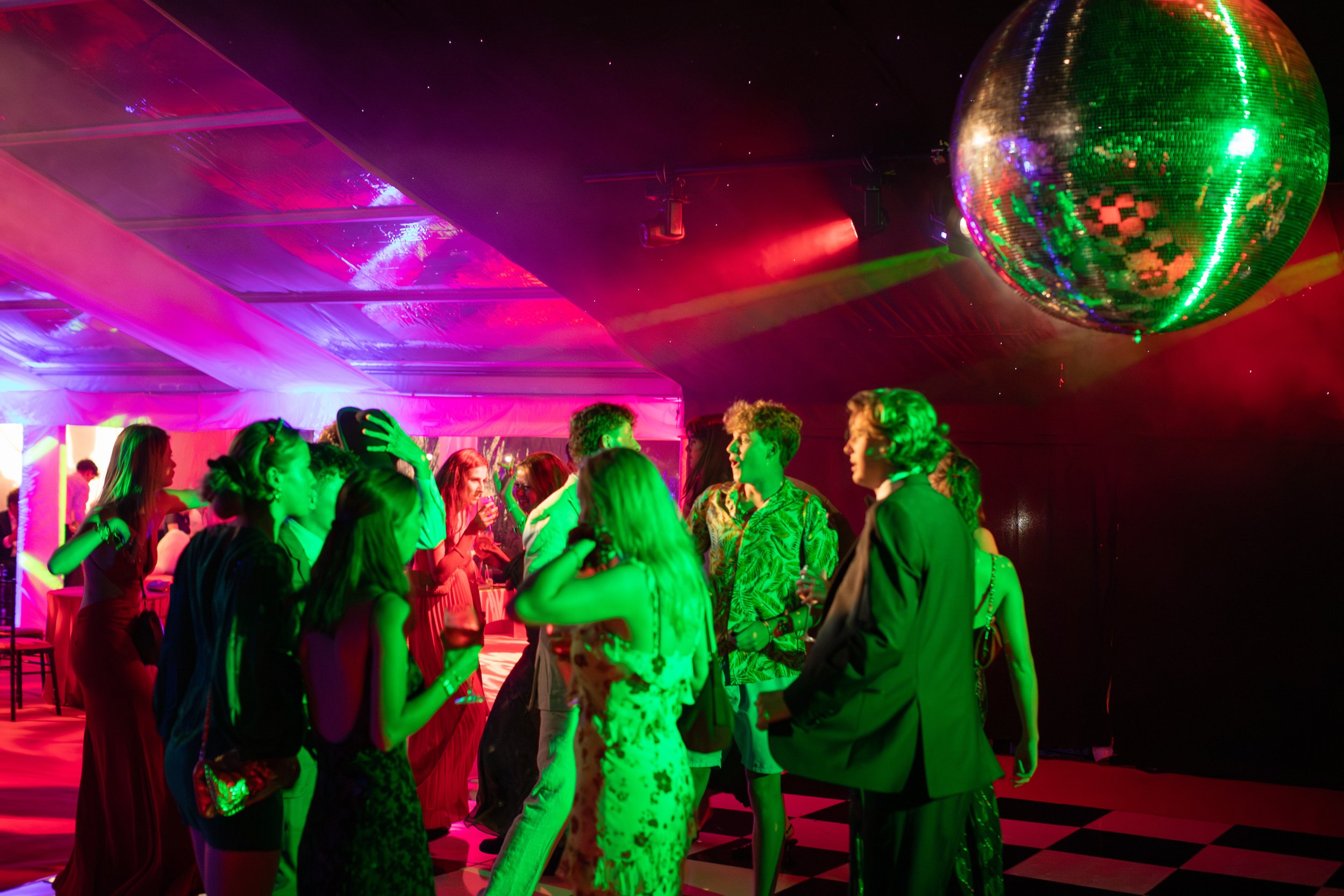 A group of people dancing and socializing at a party with colorful lighting, a large disco ball, and a checkered floor.