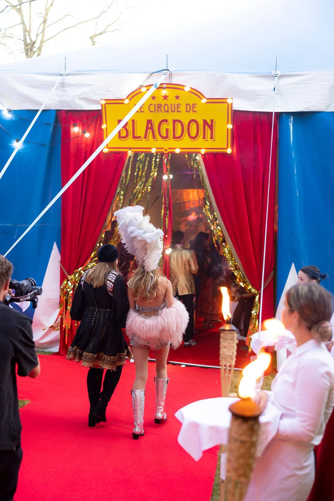 People entering a circus tent with a sign that reads 'Le Cirque de Blagd1' at an outdoor event, with red carpet, festive lights, and a woman with a feathered headpiece.