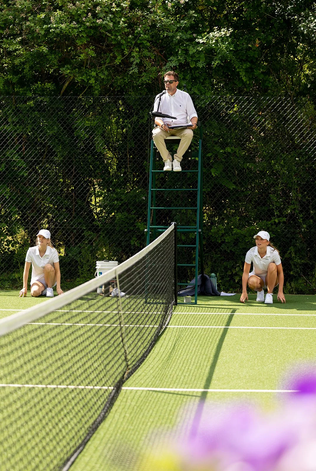 Referee sitting on a tall chair with a microphone and papers, observing a tennis match. Two female players crouching near the net, ready for a rally. A green hedge and fence in the background.
