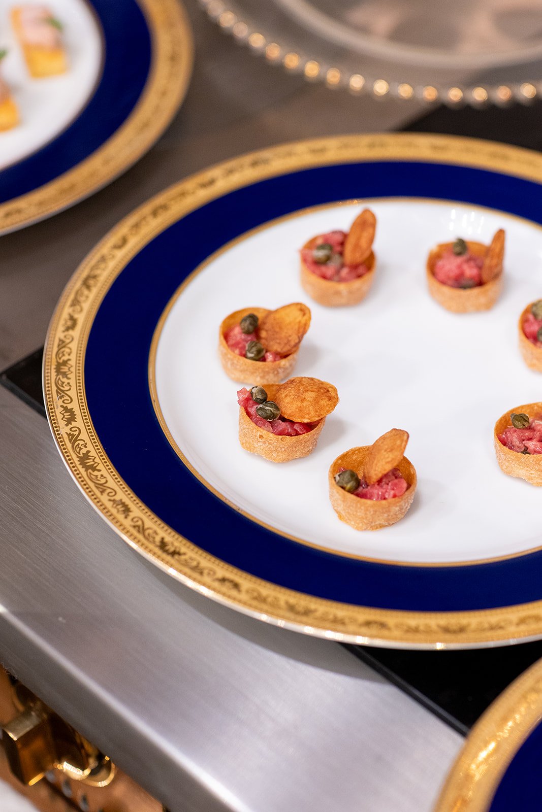 Small appetizers on a white plate with a gold and blue rim, featuring tart-like bites with pink filling, small green garnishes, and crispy chips.