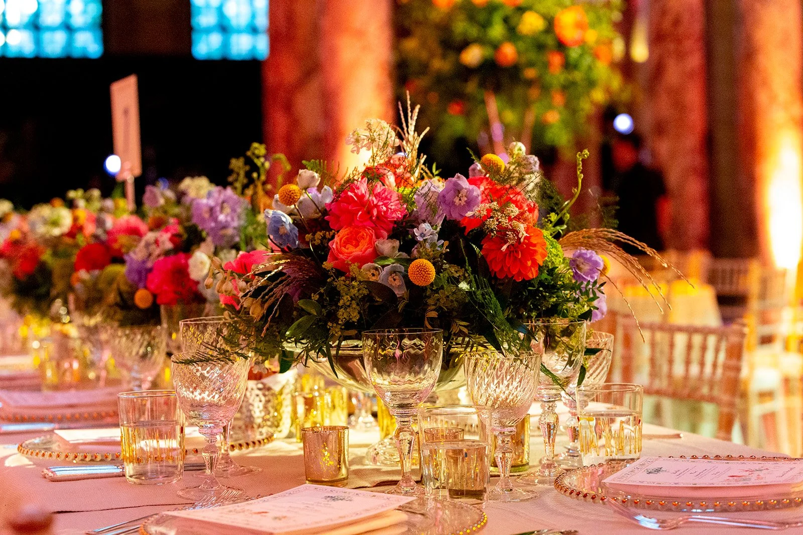 A close-up of a floral centerpiece on a dining table, with wine glasses and place settings, in a warmly lit event space with exposed brick walls and decorated with string lights.