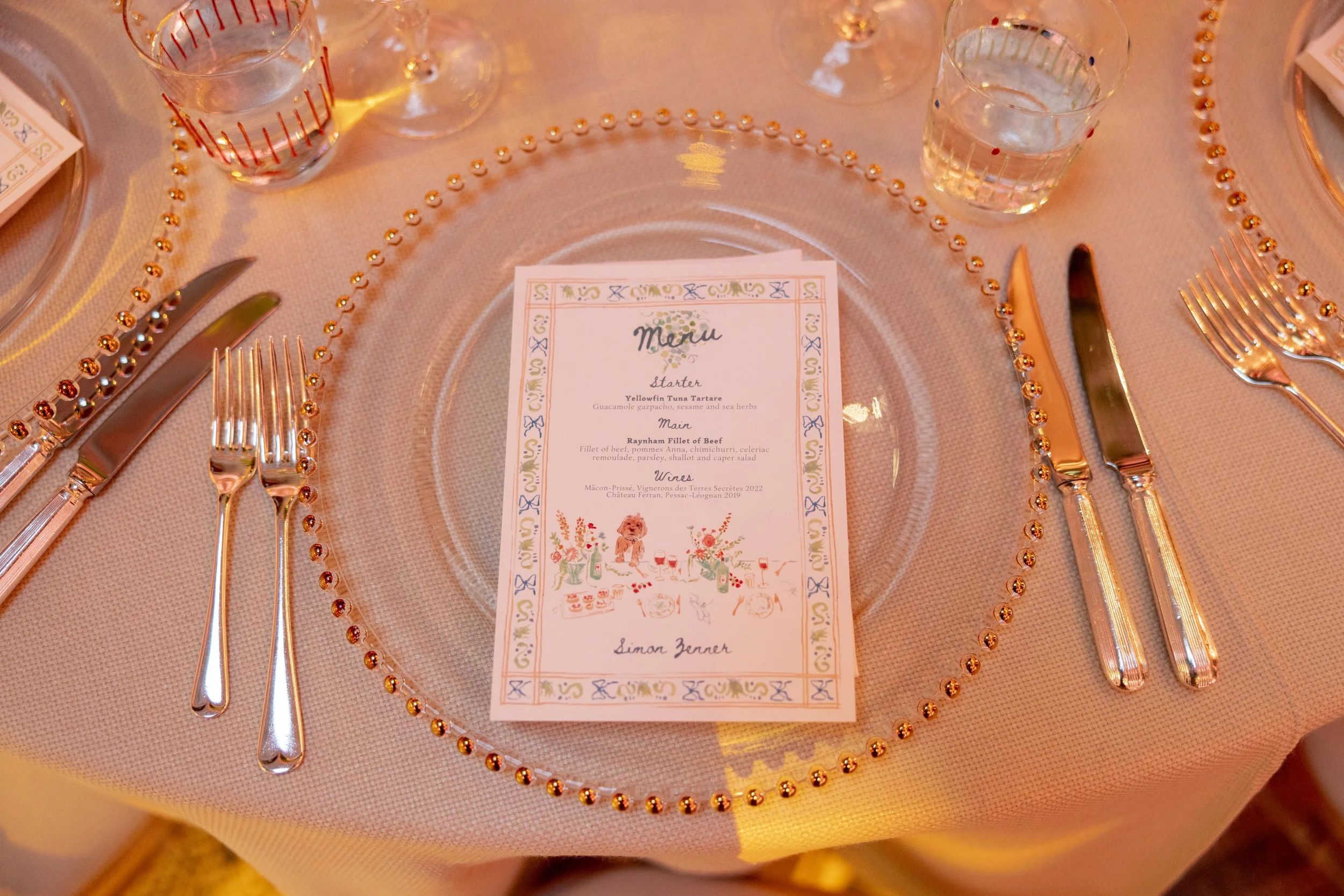 A table setting with a clear decorative plate, several forks and knives arranged symmetrically, a glass with a pattern, and a printed menu placed on the plate. The tablecloth is pink, and the overall decor is elegant and festive.