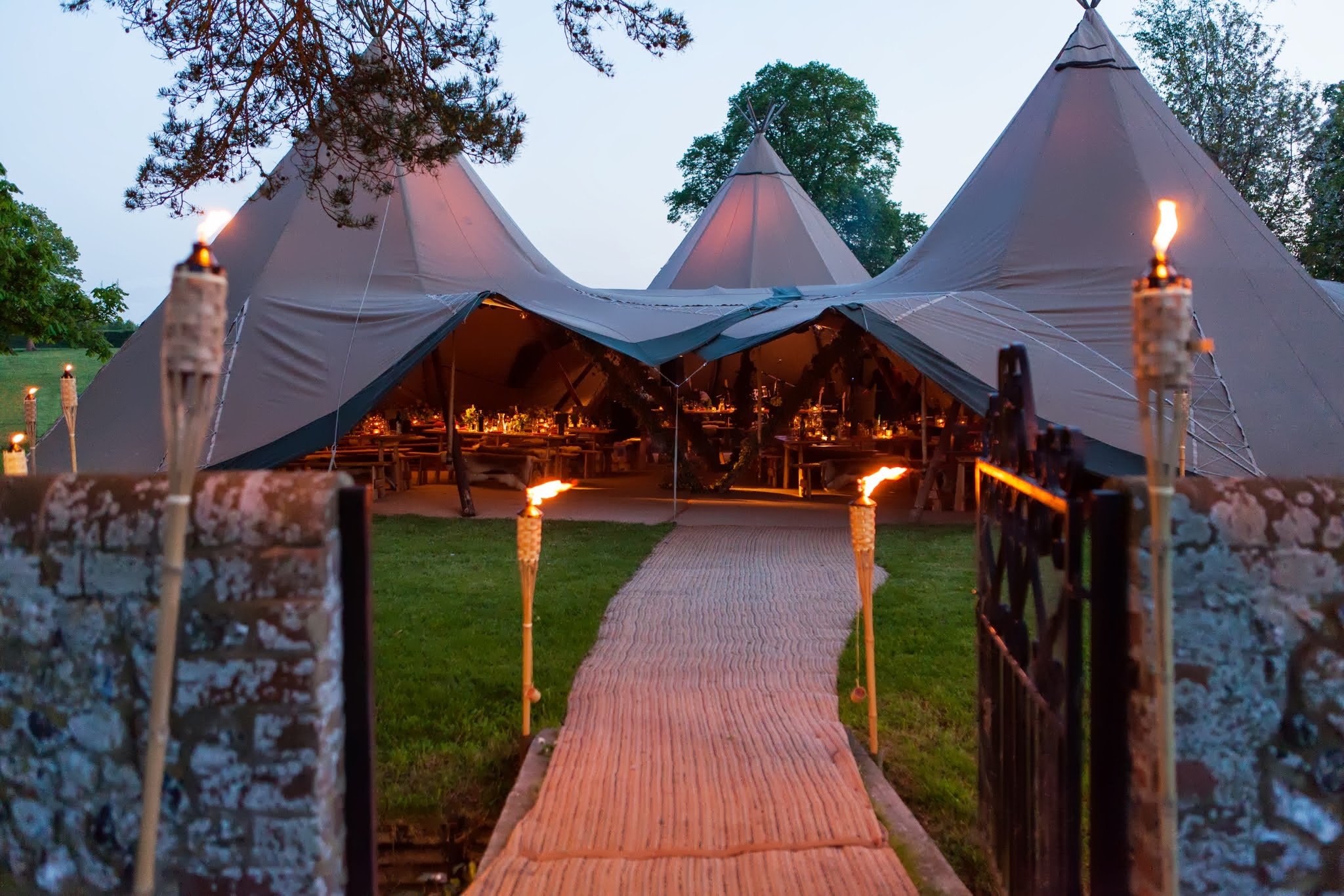 A large outdoor tent with multiple peaks, illuminated from inside, set up in a grassy area during early evening, decorated with tiki torches along a brick pathway leading to it.