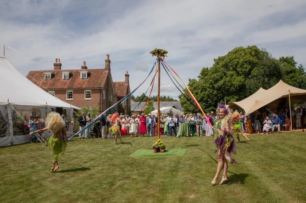 People at an outdoor garden party watching a maypole dance, with dancers in fairy costumes, tents, and a large brick house in the background.