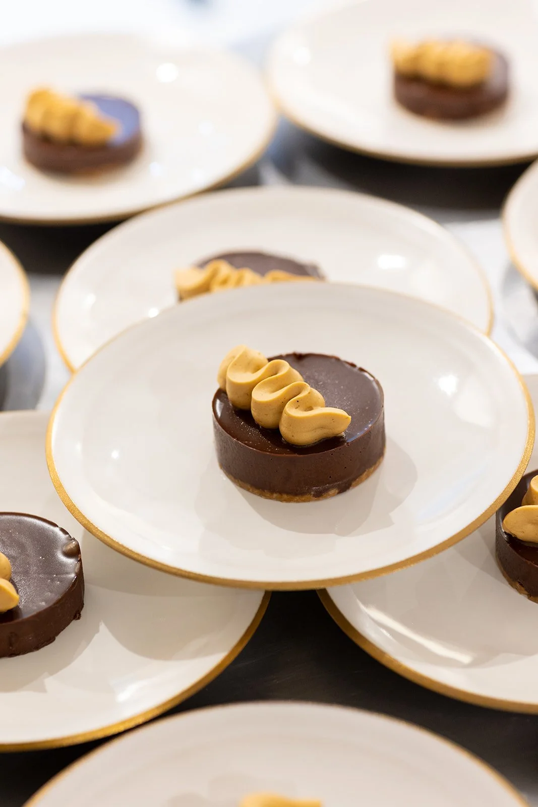 Multiple white plates with a gold rim, each holding a chocolate dessert topped with a decorative swirl of light brown cream.