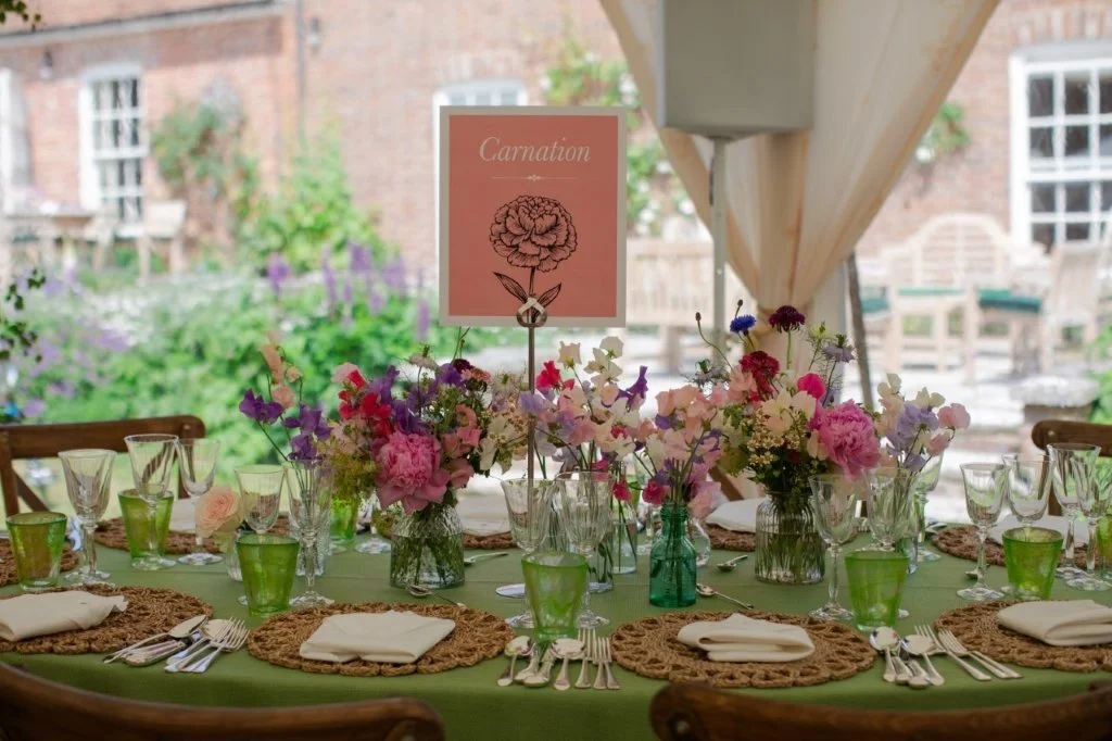 A beautifully set outdoor dining table with floral centerpieces, green glassware, and beige napkins for a wedding or special event.