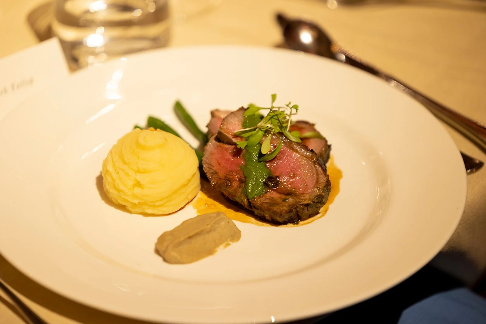 A white plate with a serving of six medium-rare steak slices topped with greens, a scoop of mashed potatoes, a dollop of brown sauce, and green beans, set on a wooden table with a spoon and glass of water in the background.