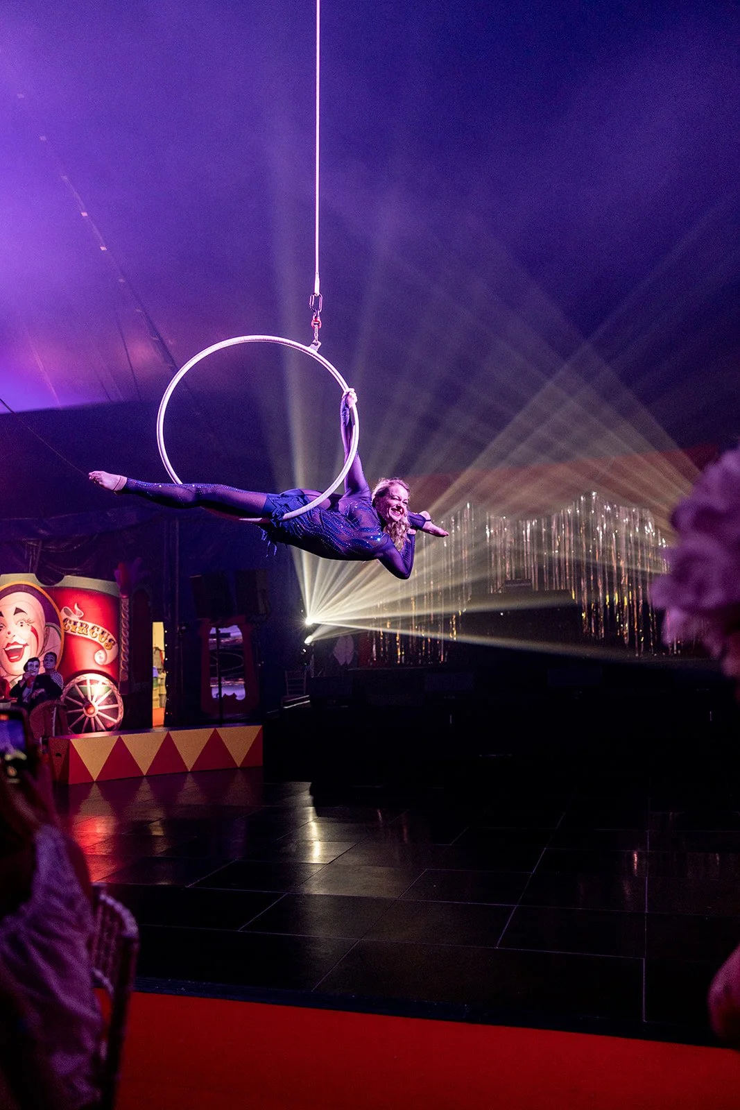 A female acrobat performing on a hoop suspended in a circus tent with colorful lights and a stage background.