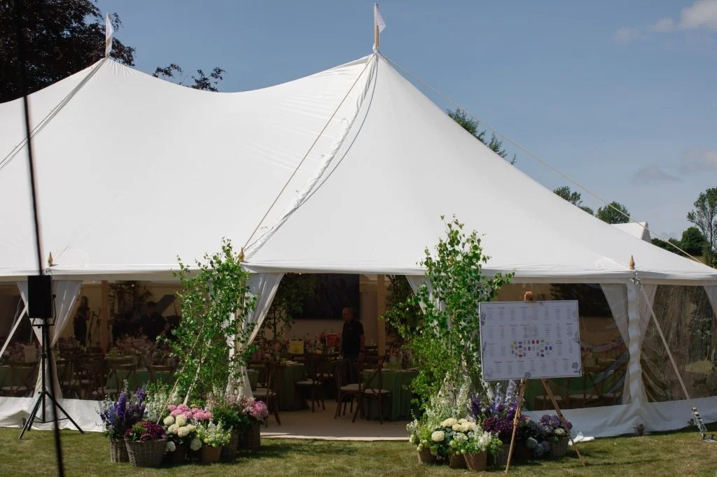 A large white event tent decorated with potted flowers and plants at the entrance, set up outdoors on a grassy area, with a few people inside preparing for an event.