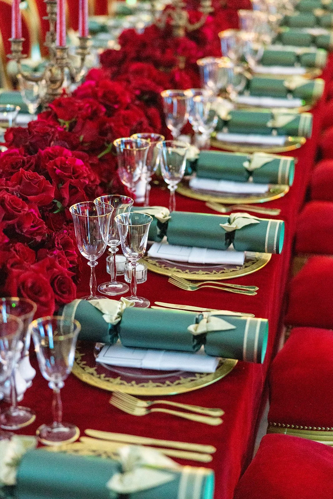 A lavish banquet table set with gold-rimmed plates, green napkins wrapped with white napkins, gold cutlery, empty wine glasses, and a centerpiece of red roses, all on a red tablecloth.