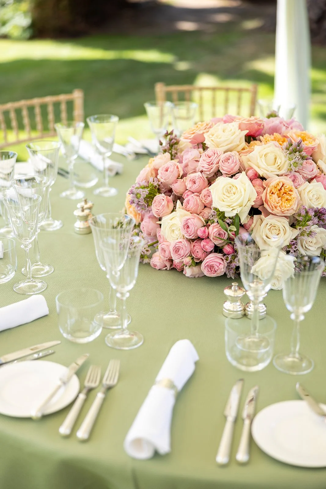 Elegant table setting with a large floral centerpiece of pink and white roses and peonies, surrounded by glassware, silverware, and white napkins, outdoors with green grass and trees in the background.