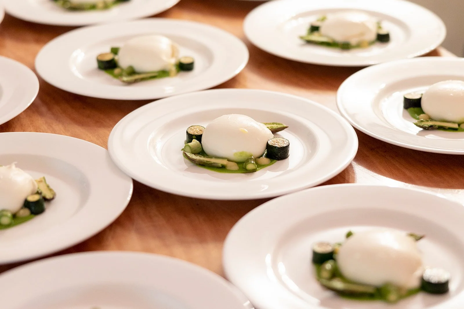 Multiple white plates each with a perfectly round poached egg, surrounded by slices of zucchini and green vegetables on a wooden table.