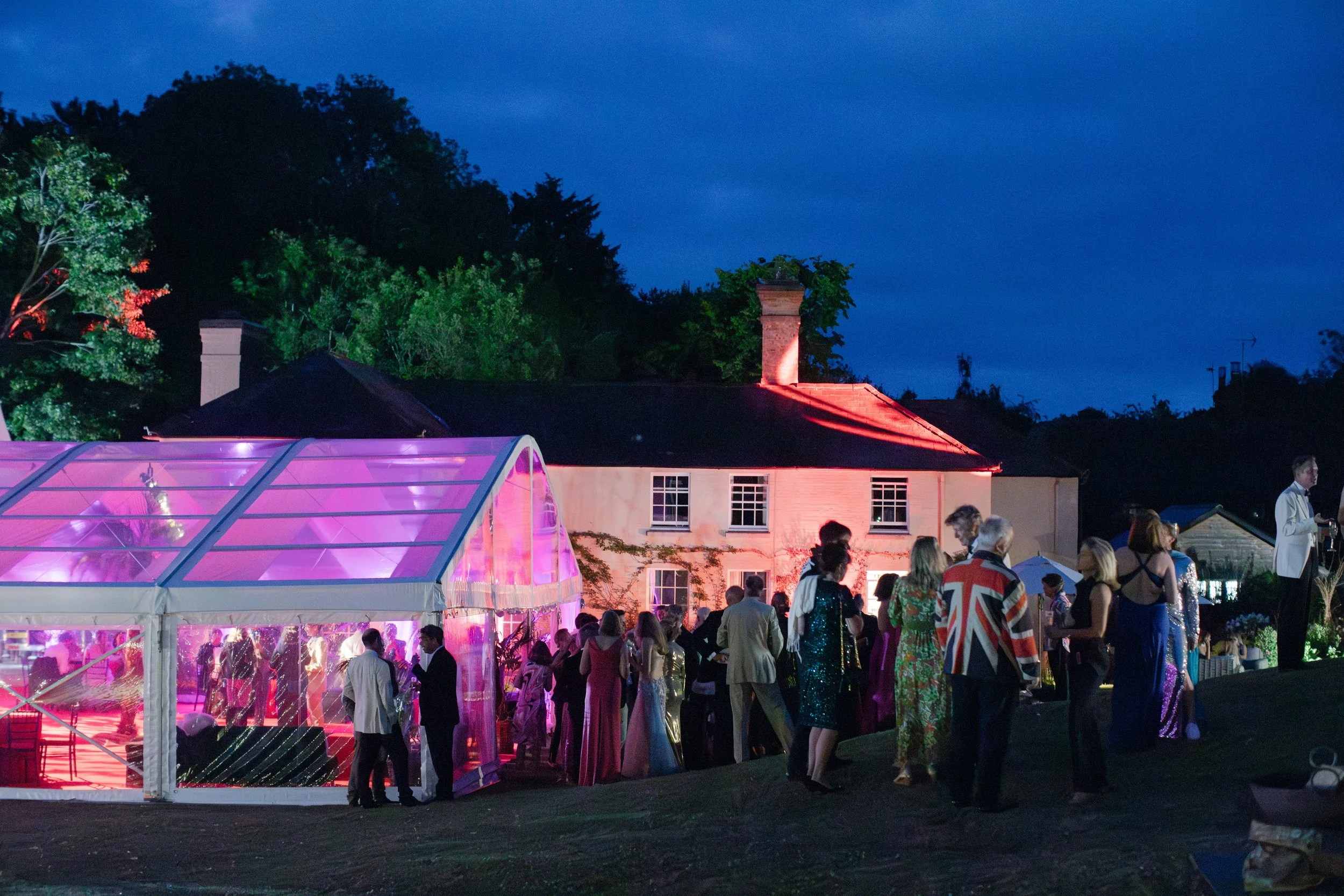 People attending an outdoor evening event near a house with illuminated windows and a pink-lit greenhouse, with trees and a dark blue sky in the background.