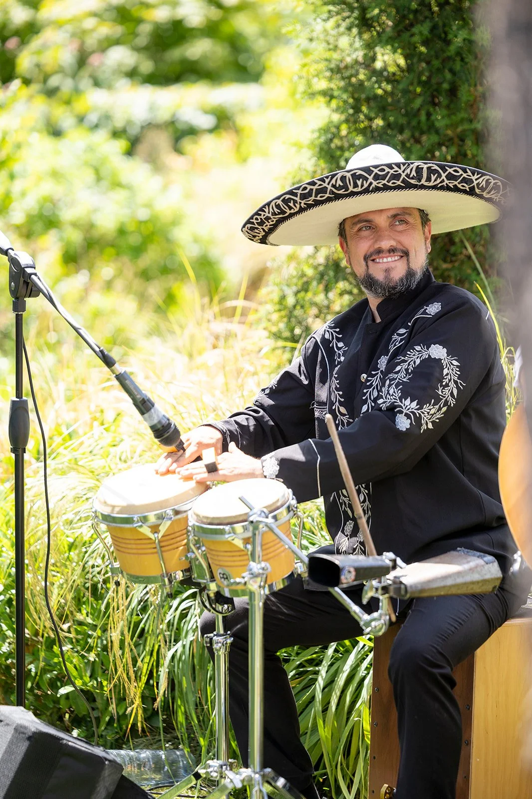 A man wearing a large sombrero with black and white embroidery, playing percussion instruments outdoors surrounded by greenery.