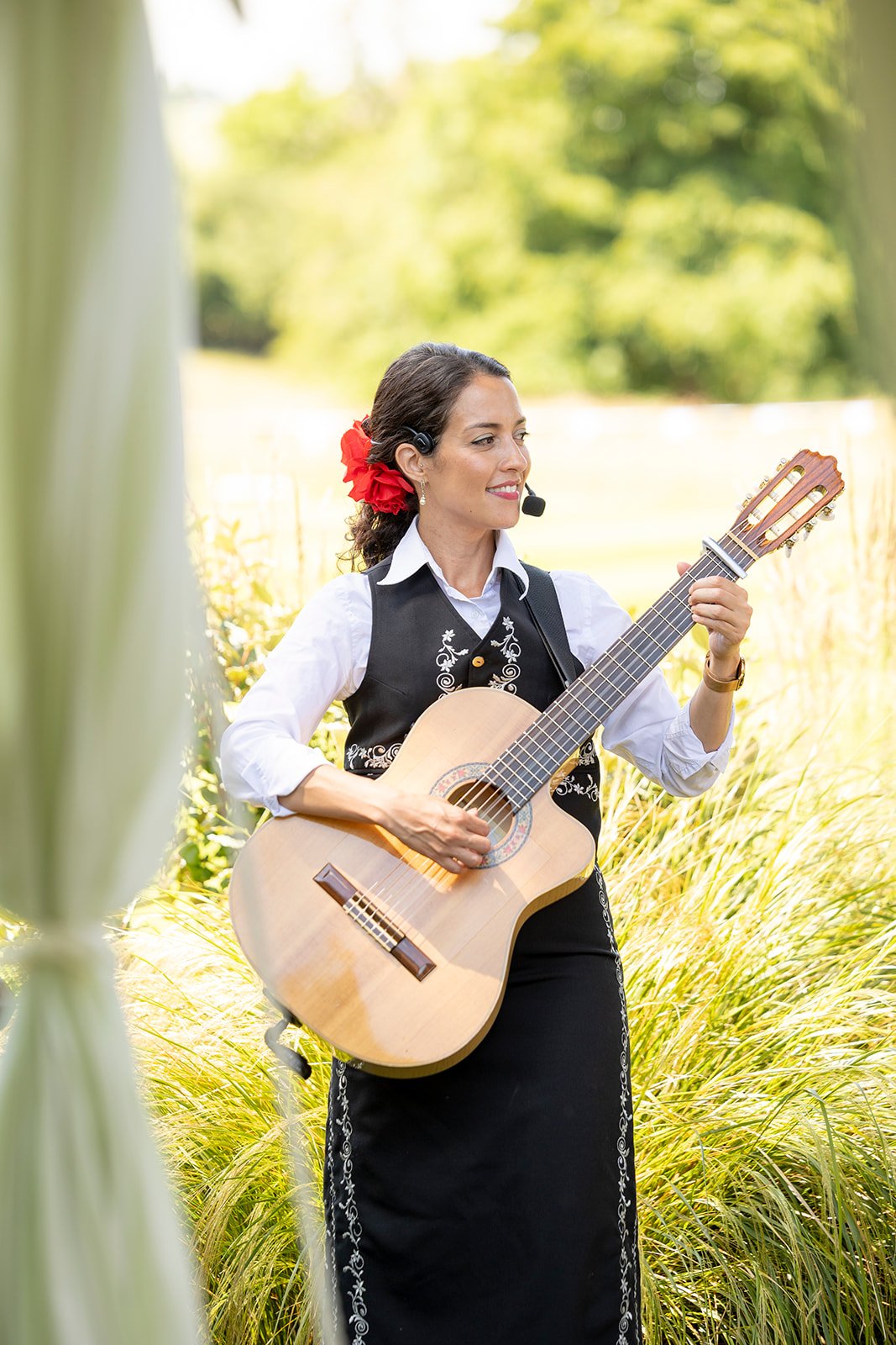 A woman wearing traditional Mexican attire, playing a guitar outdoors in a grassy, sunlit field with blurred green trees in the background, and a red flower in her hair.