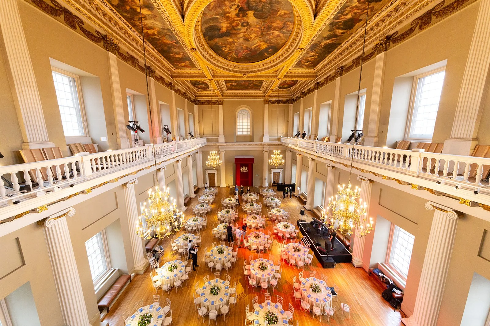 Elegant banquet hall decorated for an event with round tables covered in white tablecloths and floral centerpieces, gold chandeliers hanging from a decorated ceiling, and large windows allowing natural light. There is stage area with a piano and staf