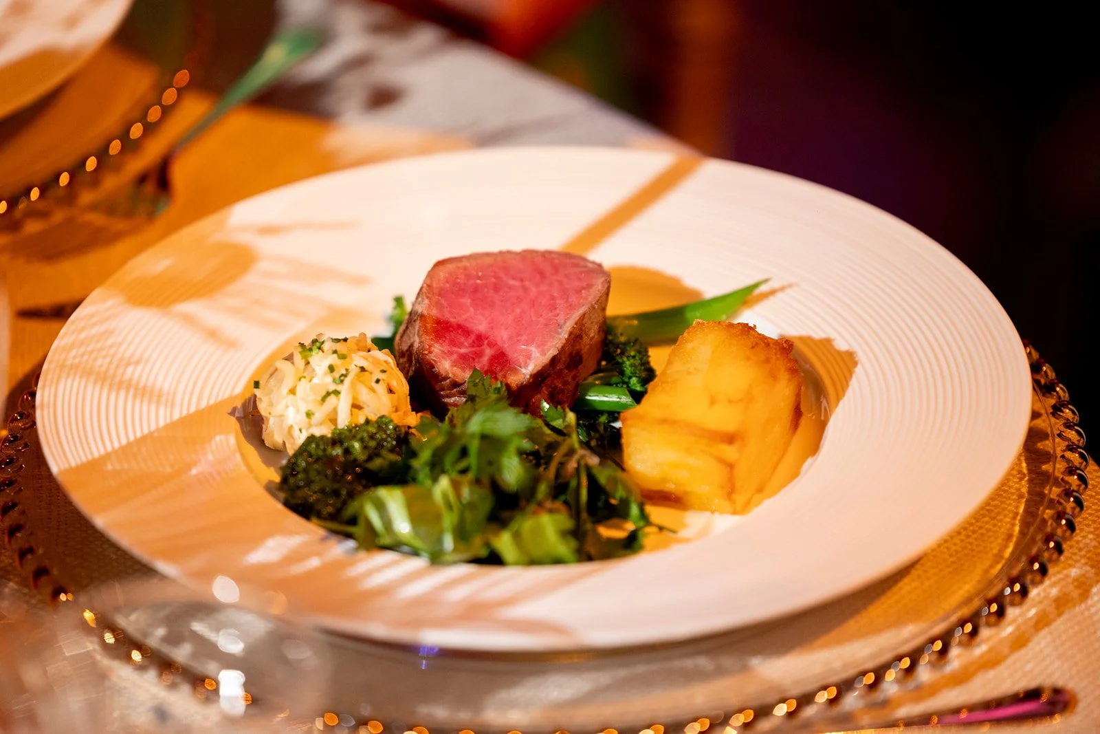 A plated dish featuring a cut of medium-rare steak, a baked potato, sautéed greens, and a portion of cole slaw, all arranged on a white plate with a decorative gold rim.