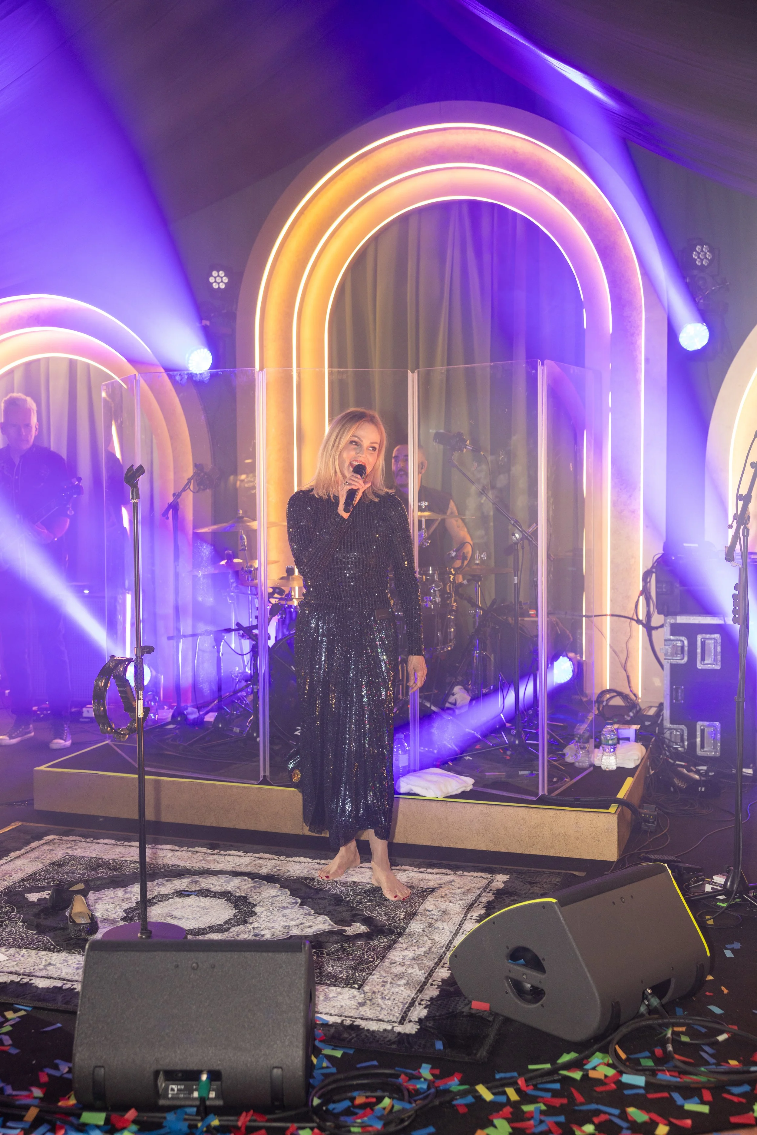 A woman in a black sequin dress is singing on stage with a microphone, barefoot. Behind her, there is a band with musicians playing instruments, surrounded by purple and yellow neon arches and lighting effects. The stage floor has a decorative rug an