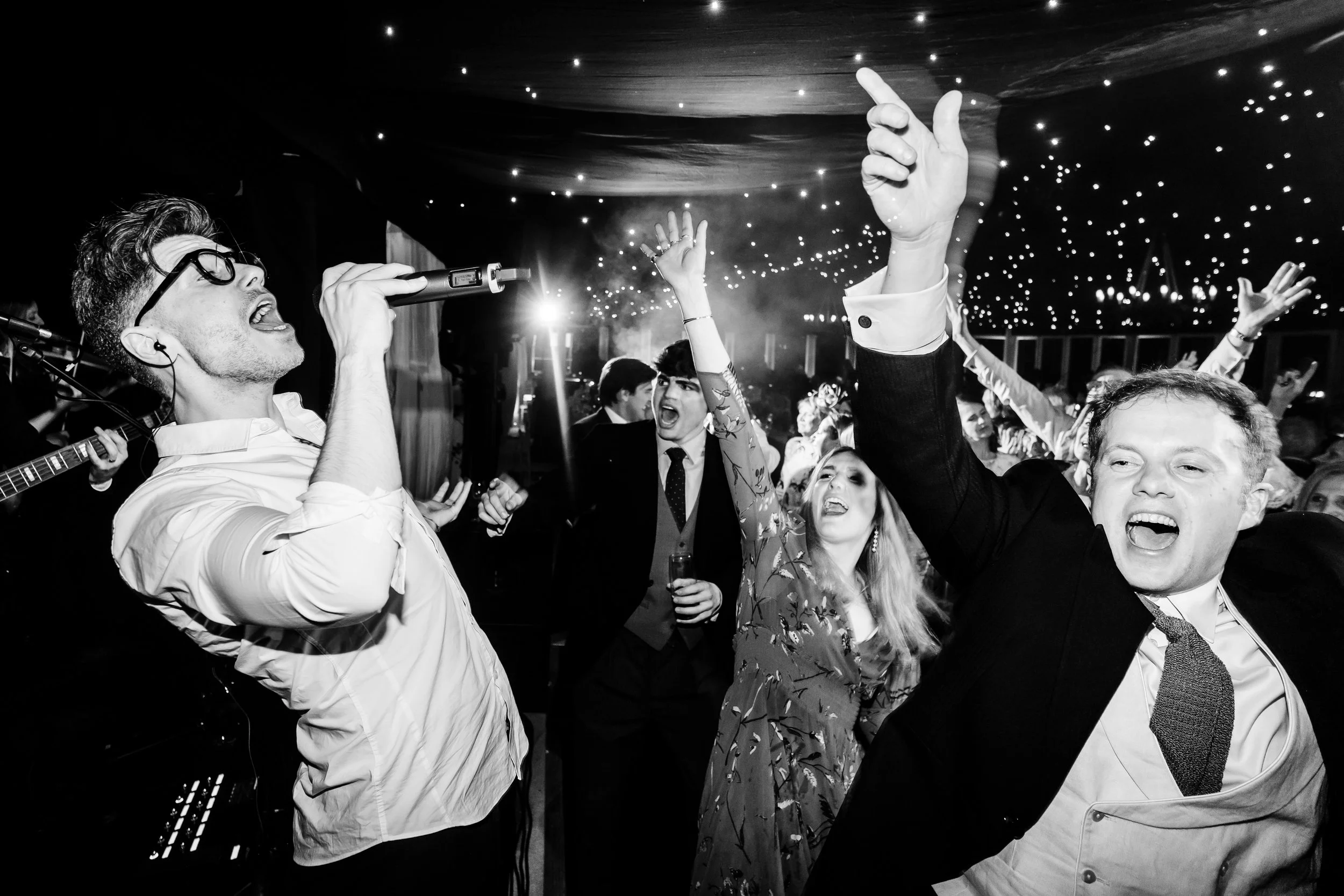People dancing and singing at a lively party or celebration, with a singer holding a microphone and the ceiling decorated with string lights.