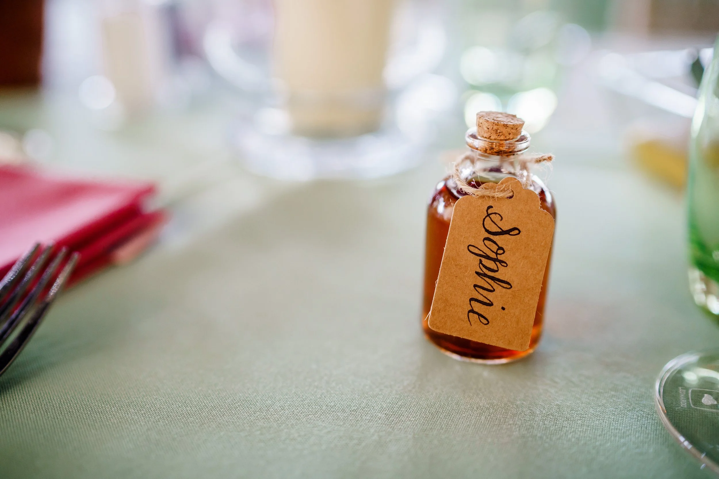 Small glass bottle with cork and a handwritten tag that says "original" attached with string, sitting on a table with blurred colorful objects in the background.