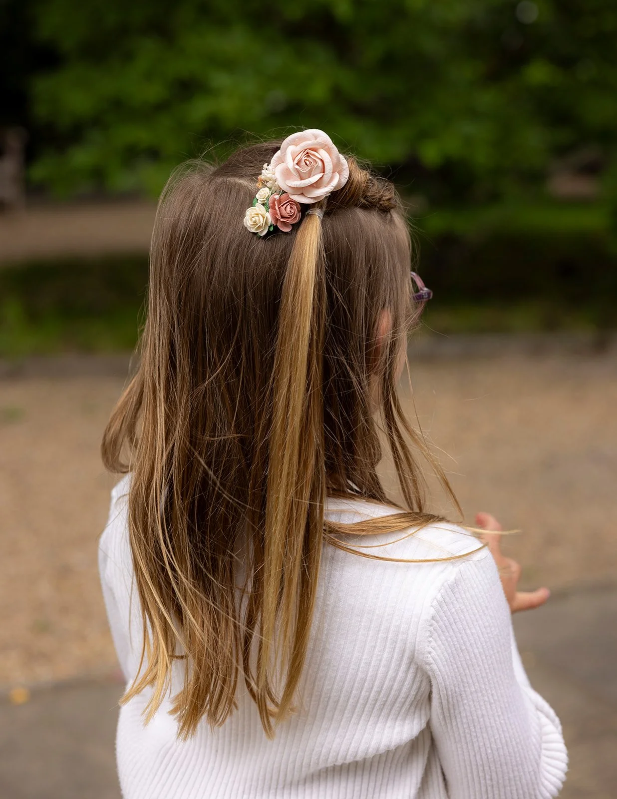 A girl with long, light brown hair wearing a floral hair accessory of pink and white roses, seen from behind outdoors.